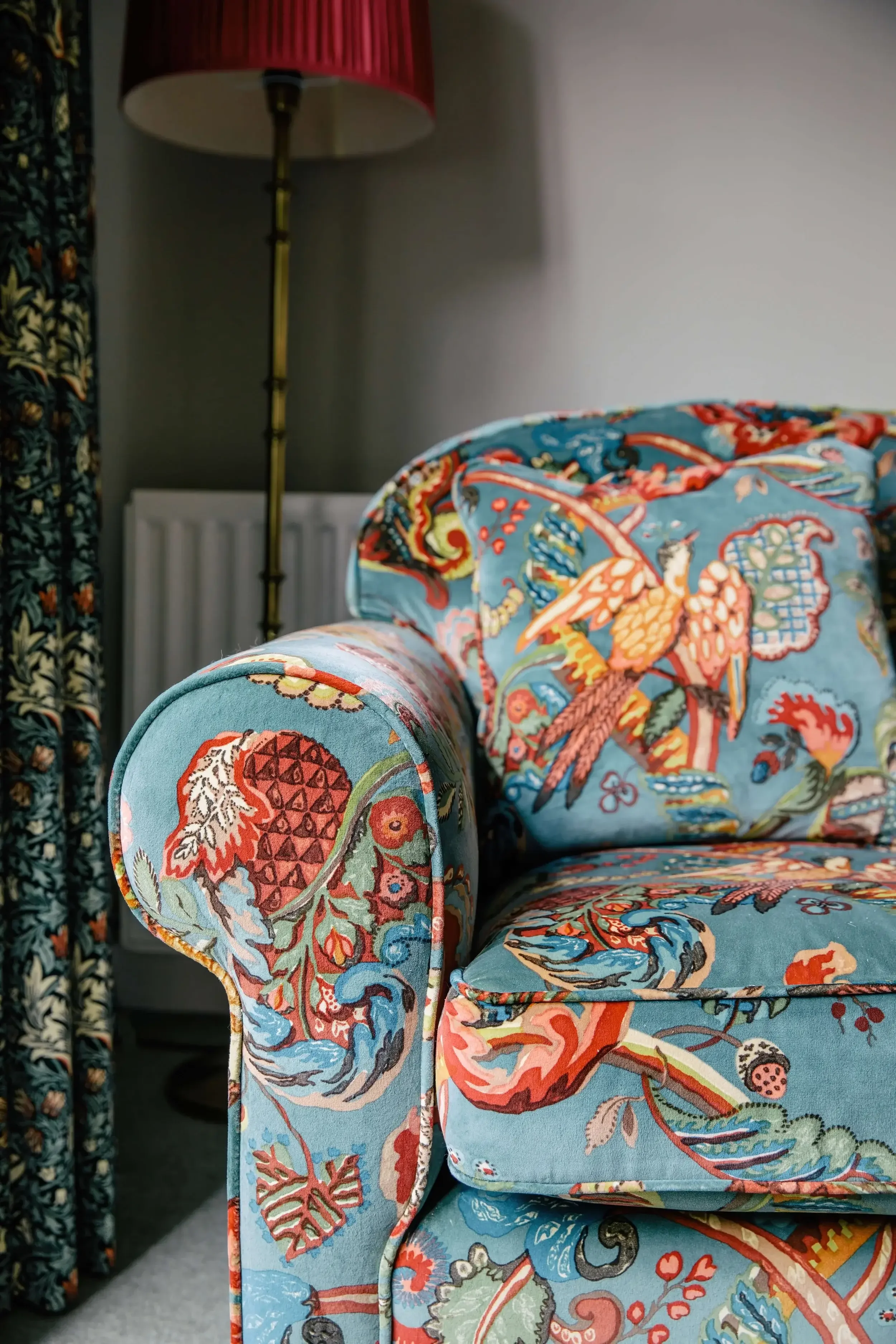 Close-up of a brightly colored upholstered armchair with a detailed floral and bird pattern, in front of a gray wall with a red-shaded floor lamp and curtains.