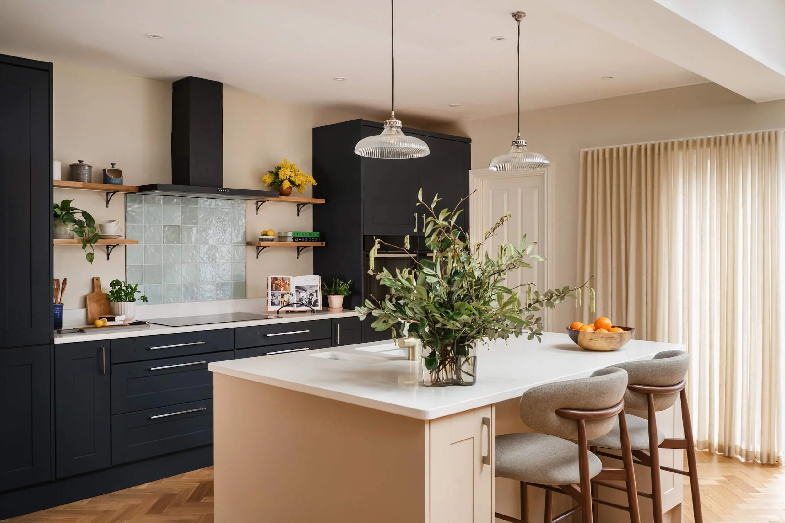 Modern kitchen with black cabinets, wooden open shelves, a white island with bar stools, a vase with green foliage on the island, and natural light from a large window with beige curtains.
