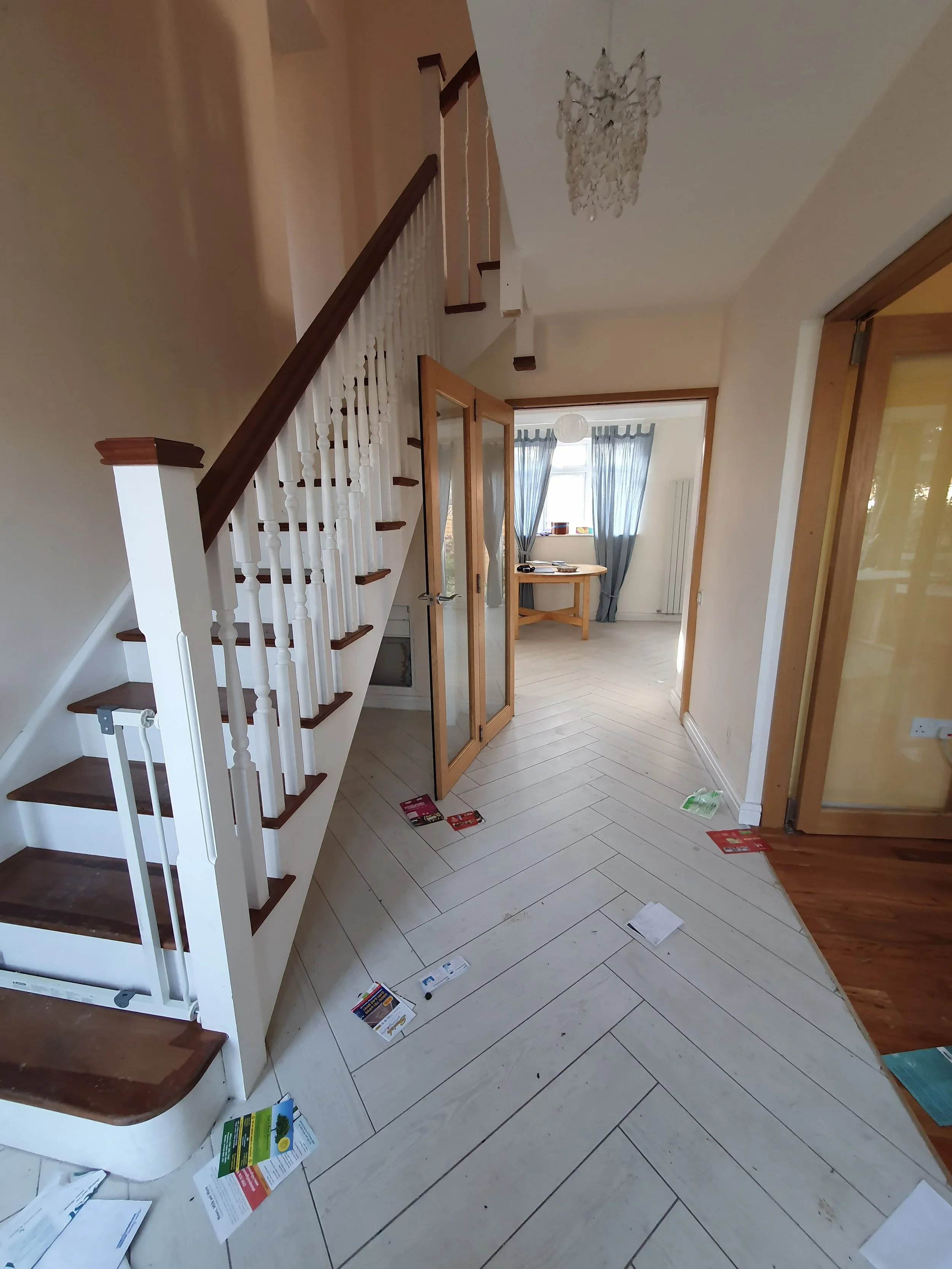 A hallway with a staircase on the left, leading upstairs. There are scattered papers and flyers on the wooden and tile floor. At the end of the hall, there is a door leading to a bright room with blue curtains and a small table.