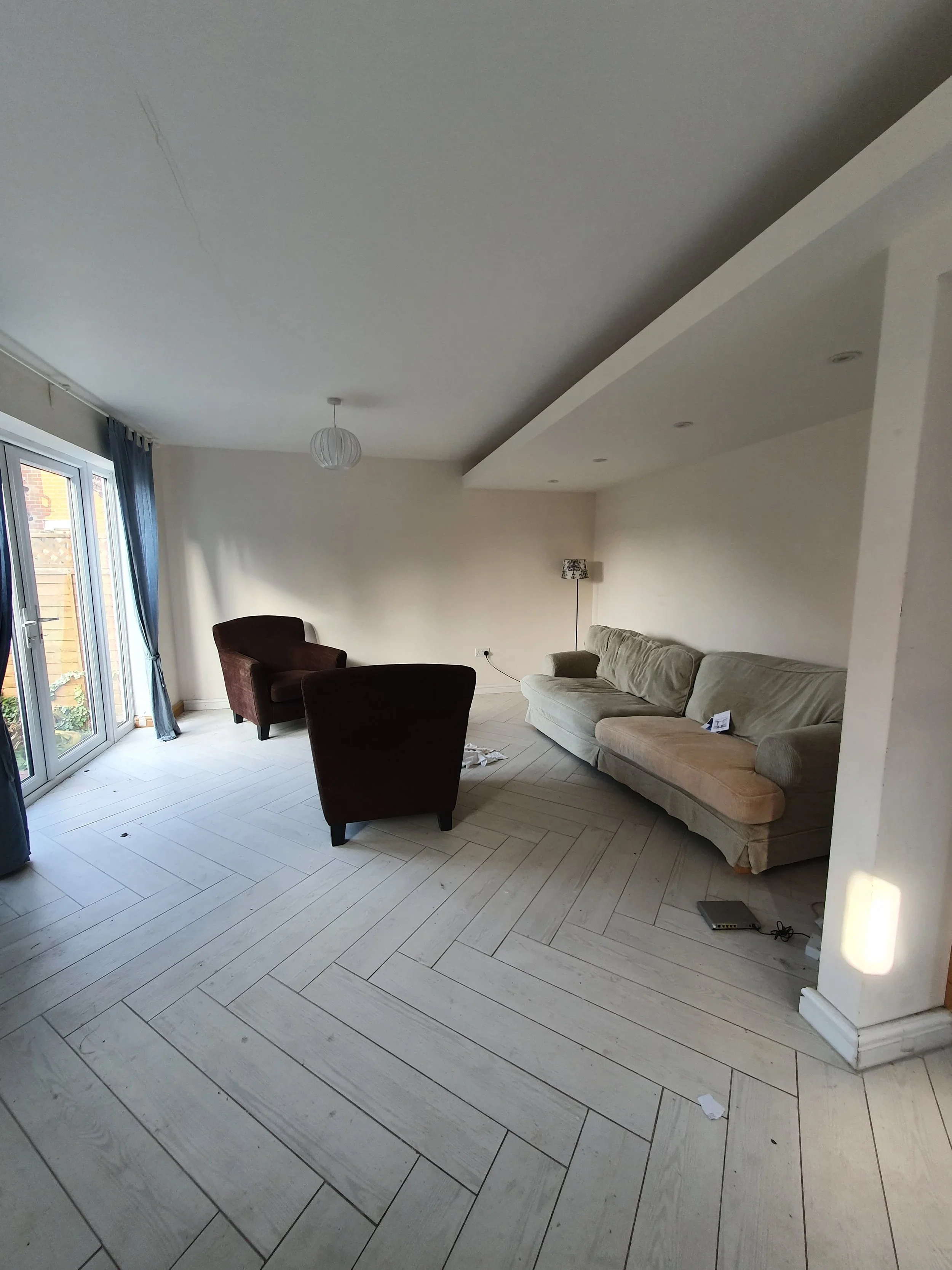 Living room with beige sofa, two dark brown armchairs, a standing lamp, and a sliding glass door with blue curtains, with some debris on the floor.