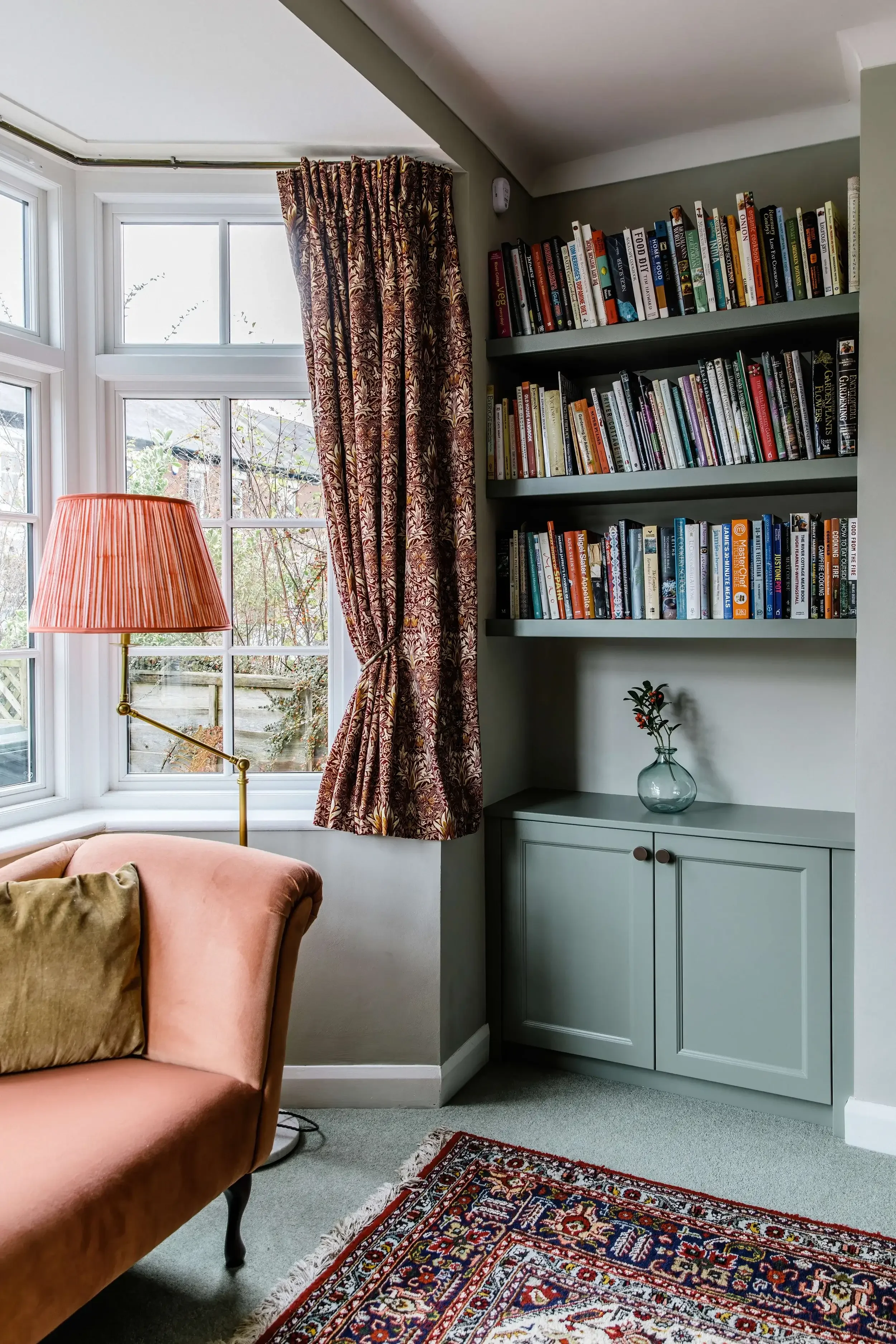 A cozy corner of a living room with a peach armchair, a rainbow-colored patterned area rug, a standing lamp with a pinkish lampshade, a white window with floral curtains, a built-in bookshelf filled with books, and a small glass vase with branches on