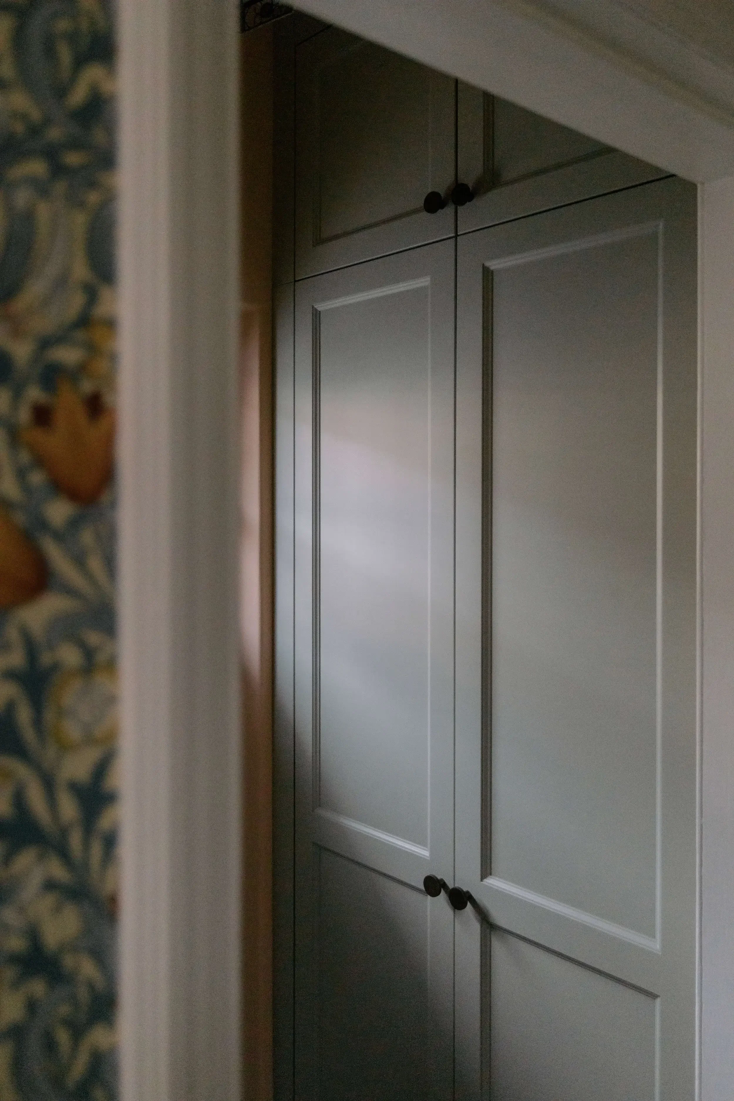 A tall, closed, gray cabinet with black knobs, viewed from a corner in a room, with a patterned wallpaper on the left side.