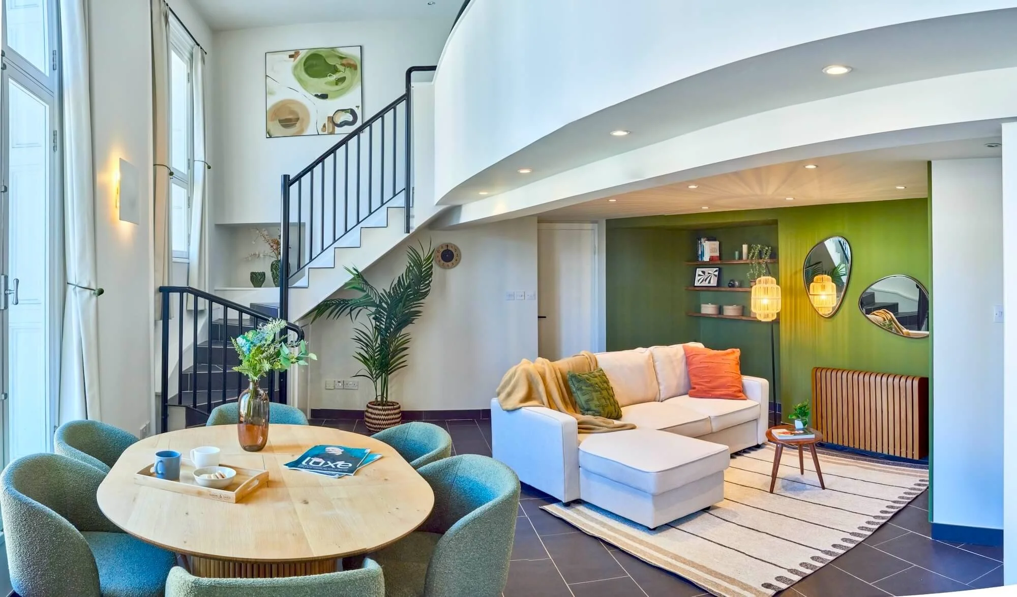 Bright living room with green accent wall, white sectional sofa with colorful cushions, striped rug, wooden side table, and built-in bookcase. Adjacent dining area has a round table with six upholstered chairs, a vase of flowers, and some dishes. Lar