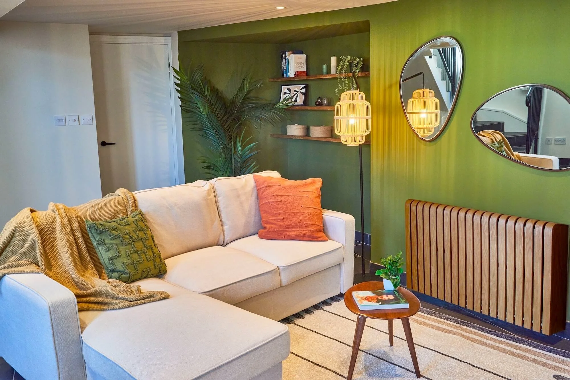 A living room with a light beige sectional sofa, decorated with green and orange pillows, a small wooden coffee table with a plant and magazine, a green accent wall with floating shelves holding books and decor, a large potted plant, two oval mirrors