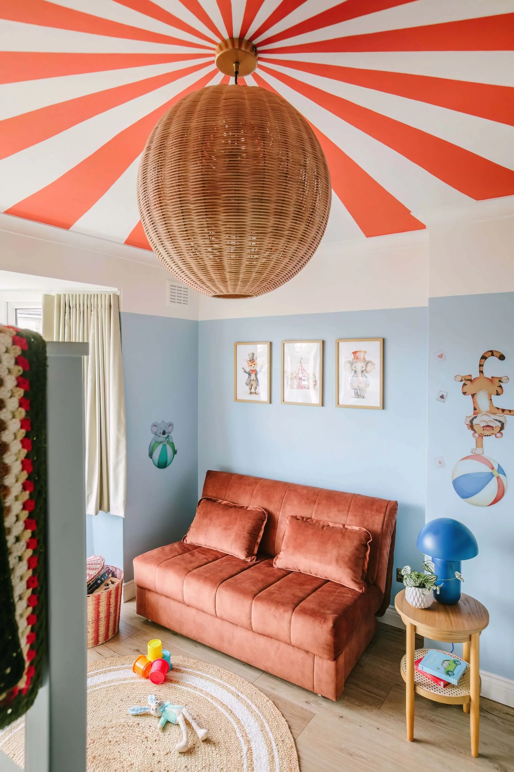 A children's room with a pink velvet sofa, a round rug, and playful cartoon wall decorations. The ceiling has a large, woven, brown pendant light and a red and white striped design radiating from the center.