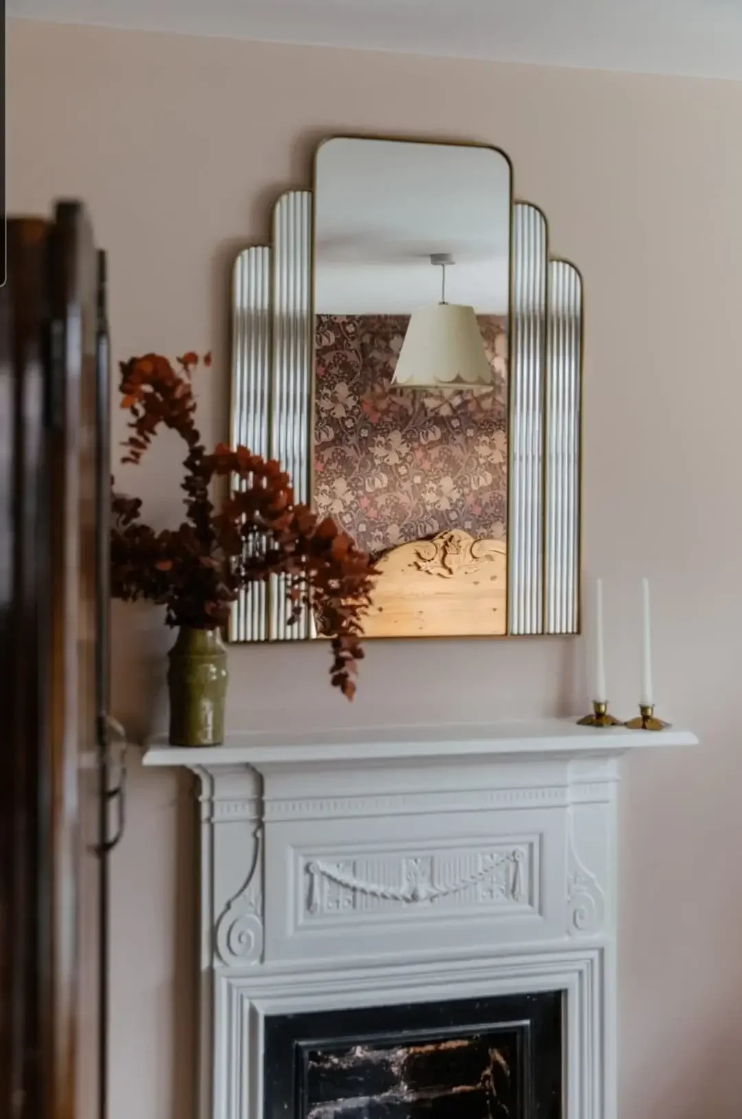 A mirror above a white fireplace mantel, reflecting a ceiling lamp and wallpapered wall, with a vase of brown flowers and two candles on the mantel.