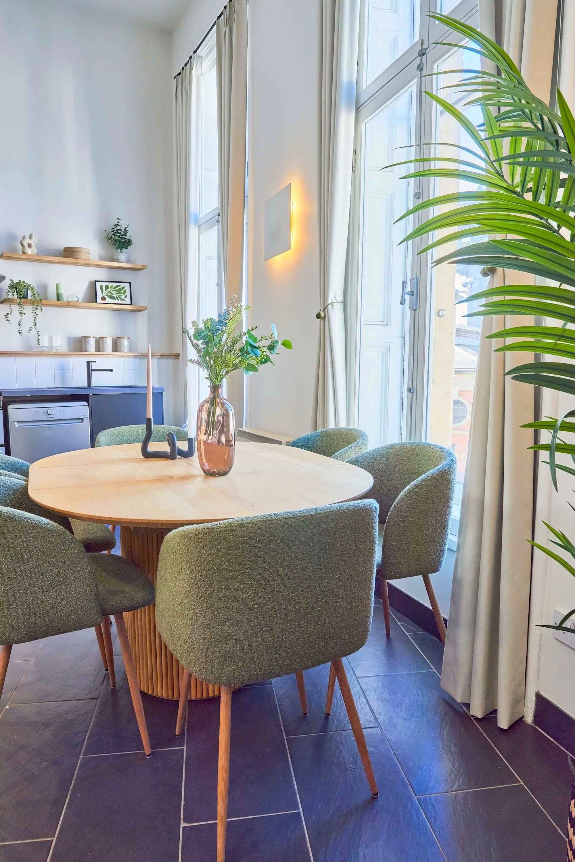 Bright dining area with a round wooden table, six green upholstered chairs, a vase with greenery, and natural light from large windows with white curtains, decorated with minimal green and neutral decor.