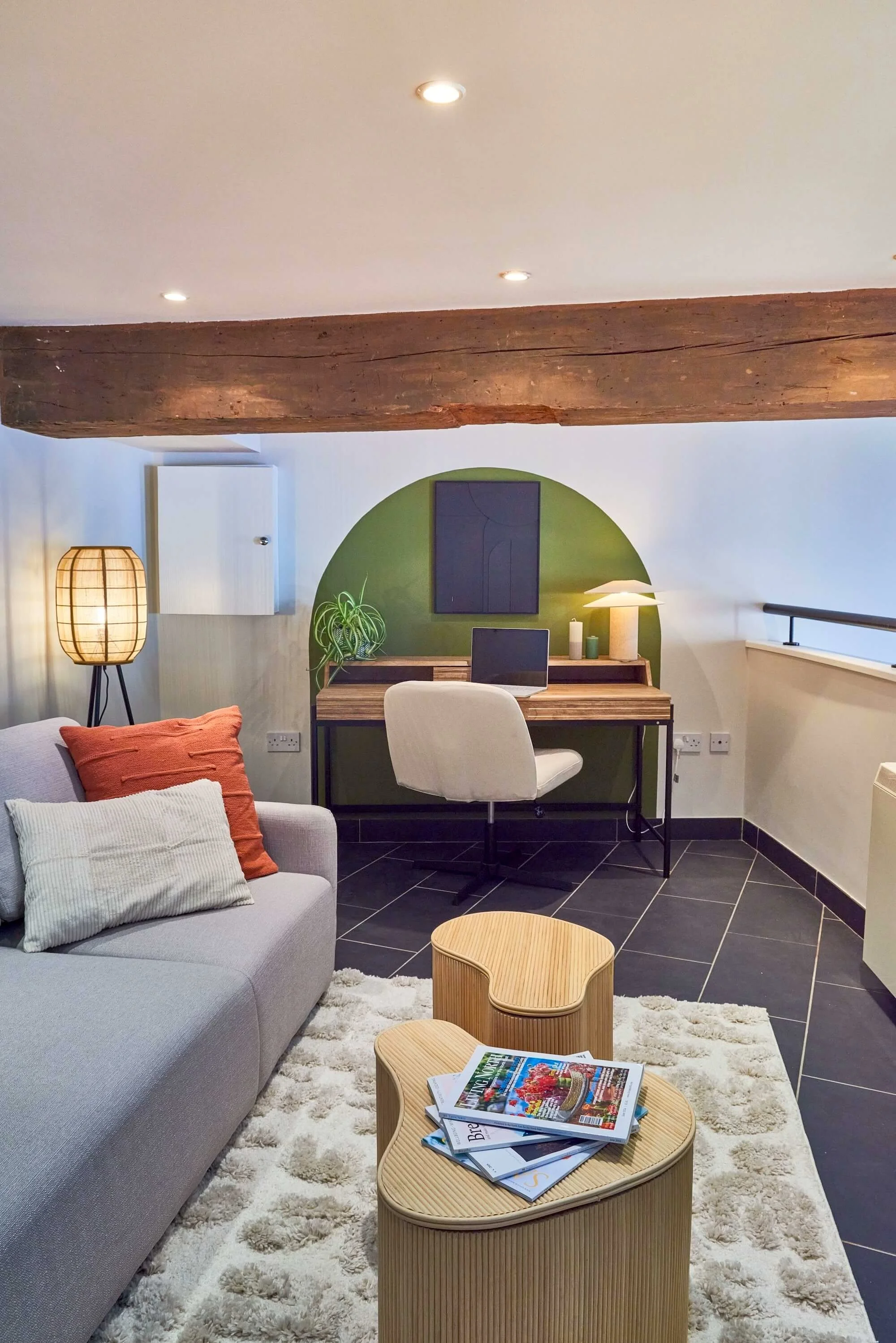 A cozy living space with a gray sofa with orange and white pillows, a round beige rug, a wooden coffee table with magazines, a desk with a computer, a beige office chair, a tall lamp, a wall lamp, a green accent wall, a wooden beam on the ceiling, an