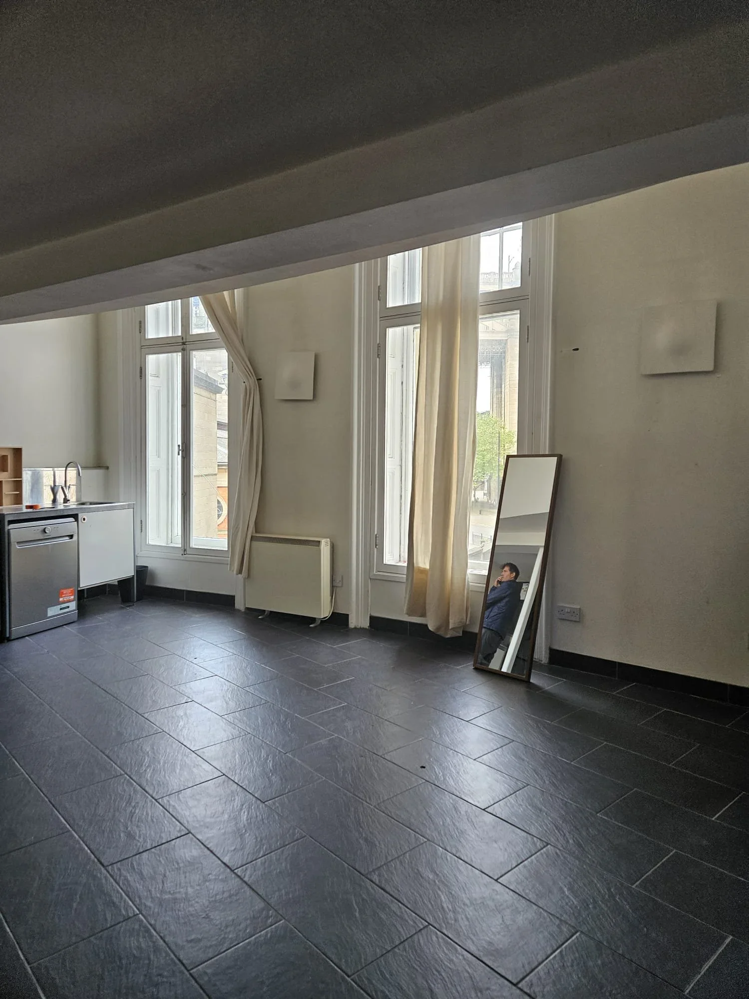 An empty room with large windows, cream-colored curtains, a radiator, a mirror leaning against the wall, and a small kitchen area with a dishwasher and a sink.
