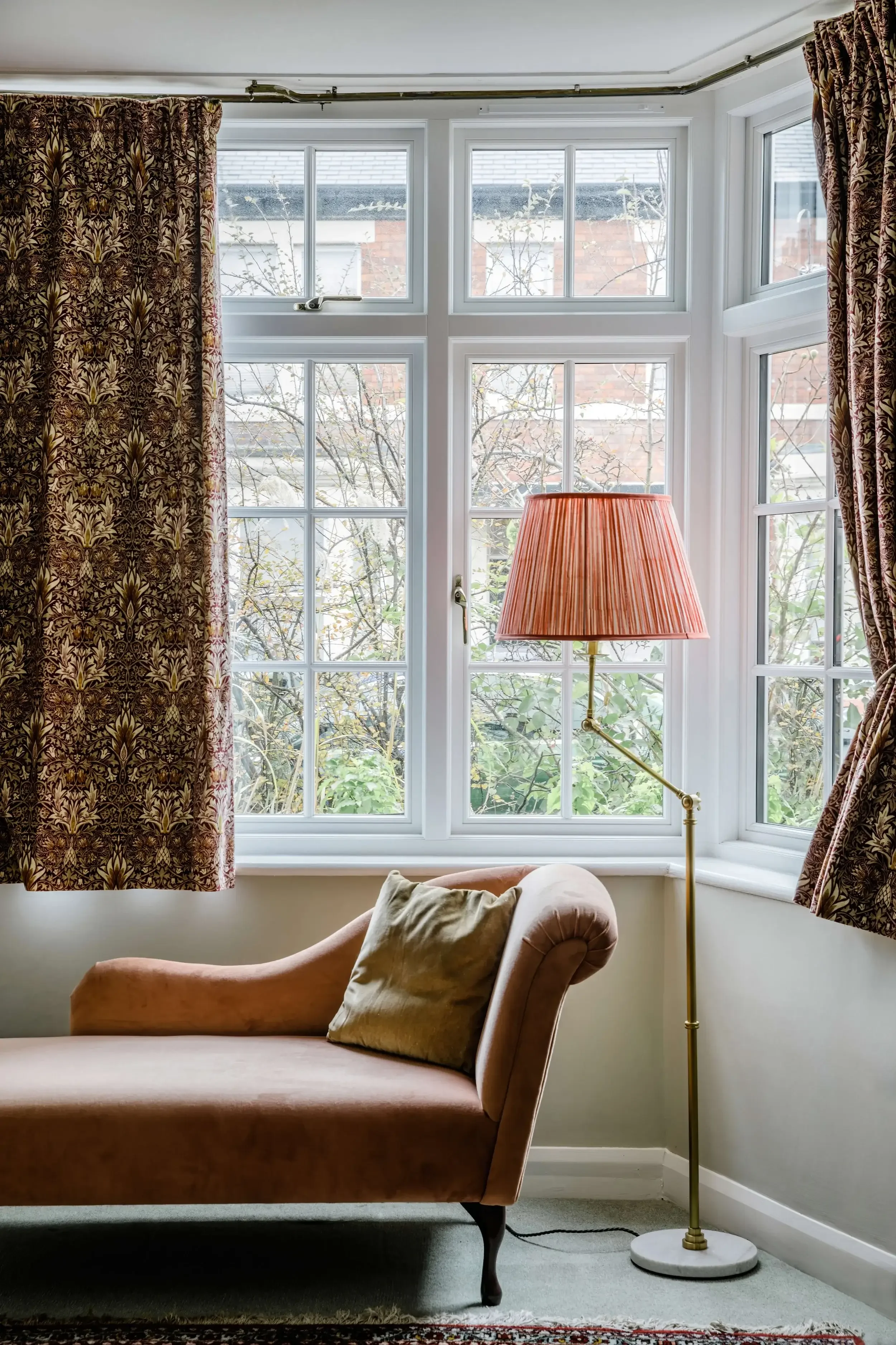 A cozy living room corner with a pink velvet chaise lounge, a beige cushion, a floor lamp with a pink pleated shade, and a large window with patterned curtains showing a view of trees outside.