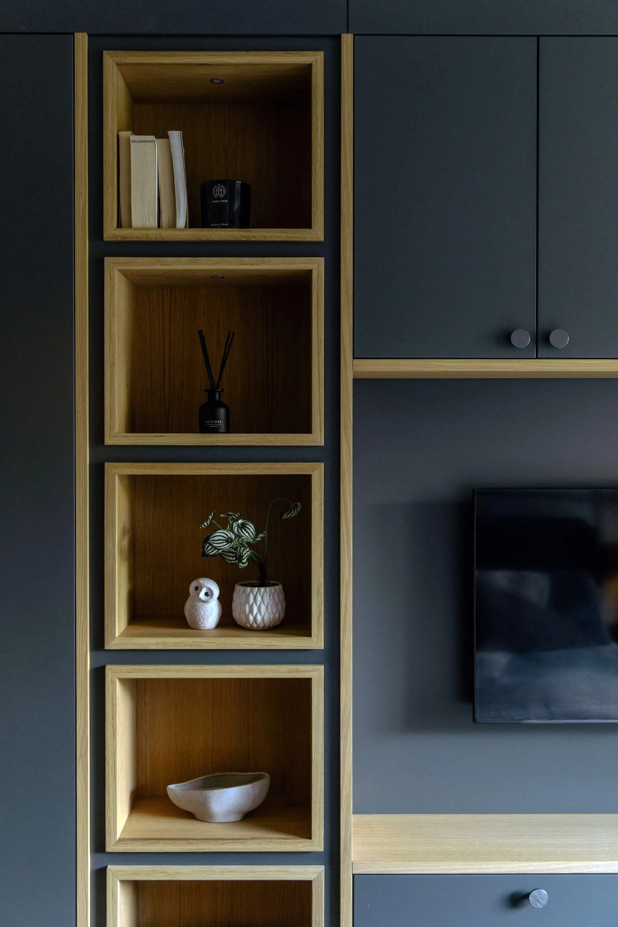 Interior home decor with wooden shelves built into a dark wall, displaying books, a small black diffuser, a white owl figurine, a sensor chip, and a ceramic bowl.