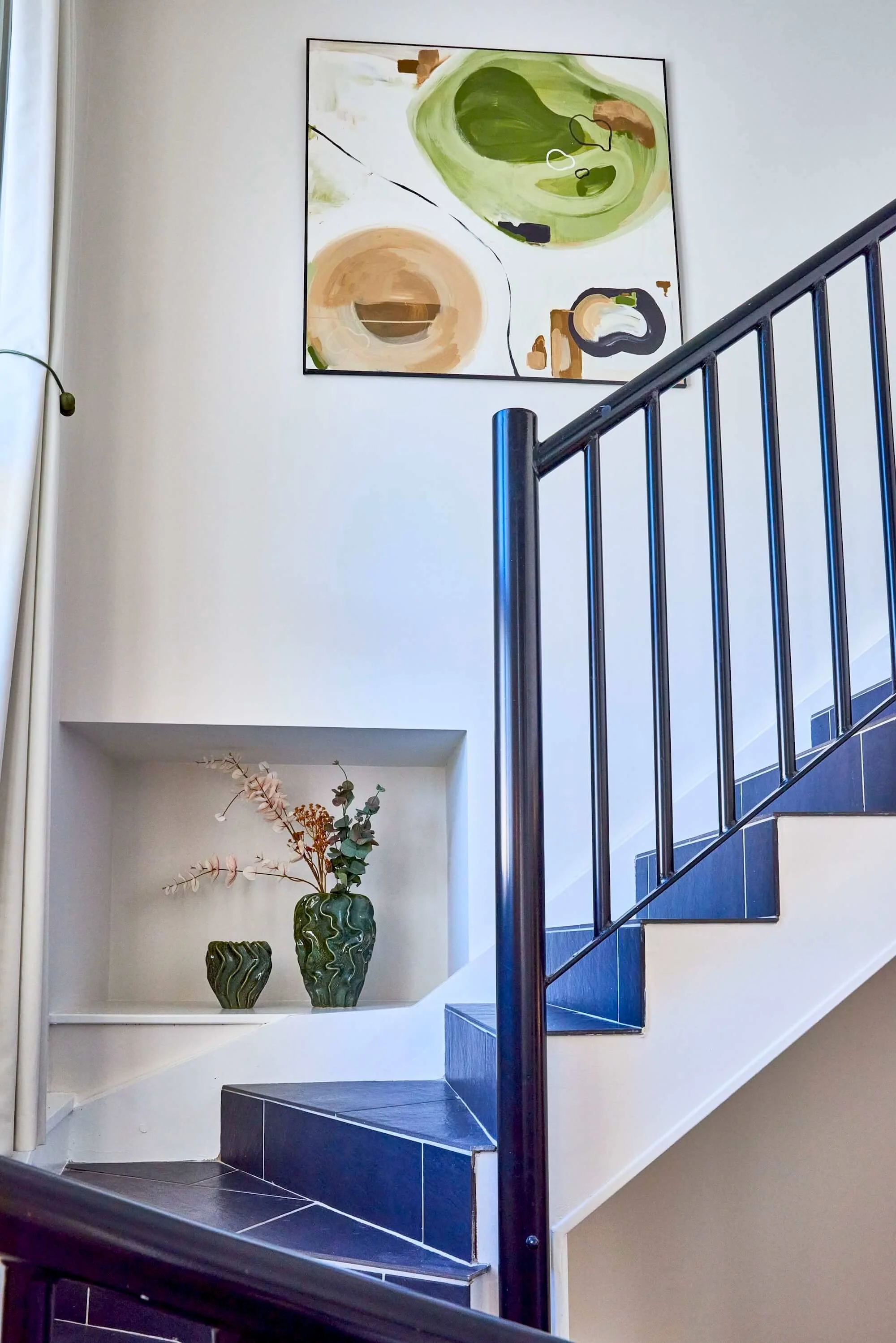 A staircase with dark blue tiles, a black metal railing, and light-colored walls. There are decorative vases with dried flowers on a shelf and a large abstract painting with green, beige, and brown shapes hanging above.