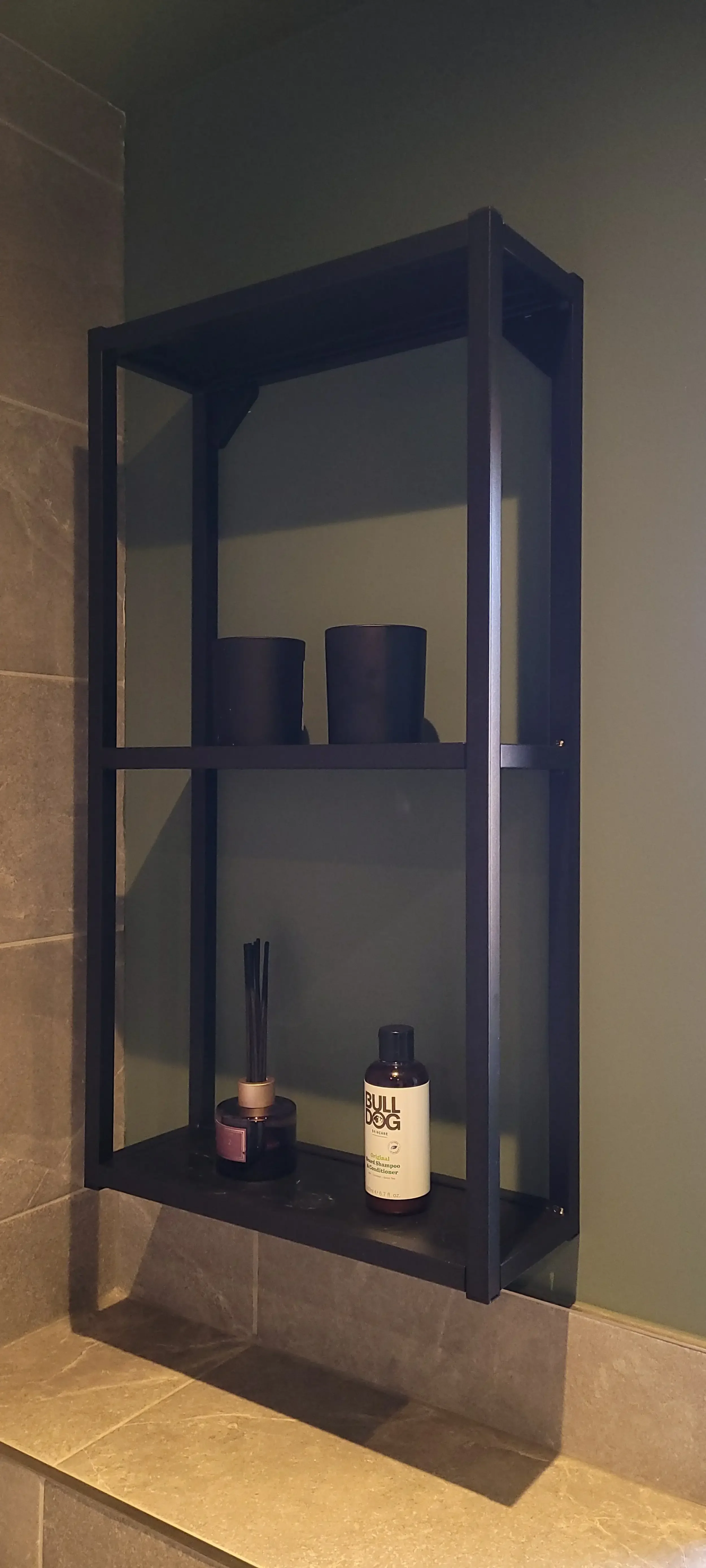 A black metal wall shelf mounted on a green wall in a bathroom. The shelf holds two black cups on the top and a black reed diffuser and a bottle of BULLDOG shampoo on the bottom.