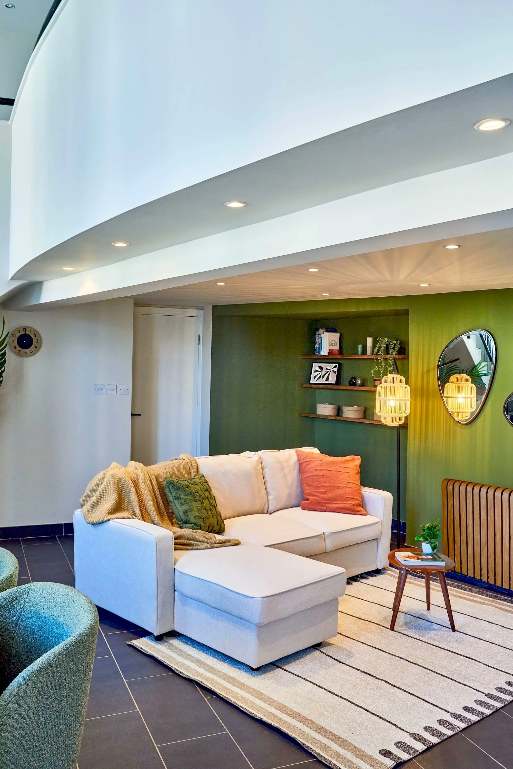 Living room with white sectional sofa, green and orange pillows, wooden side table with plant, green accent wall with wooden shelves, round mirrors, and wicker pendant lamps.