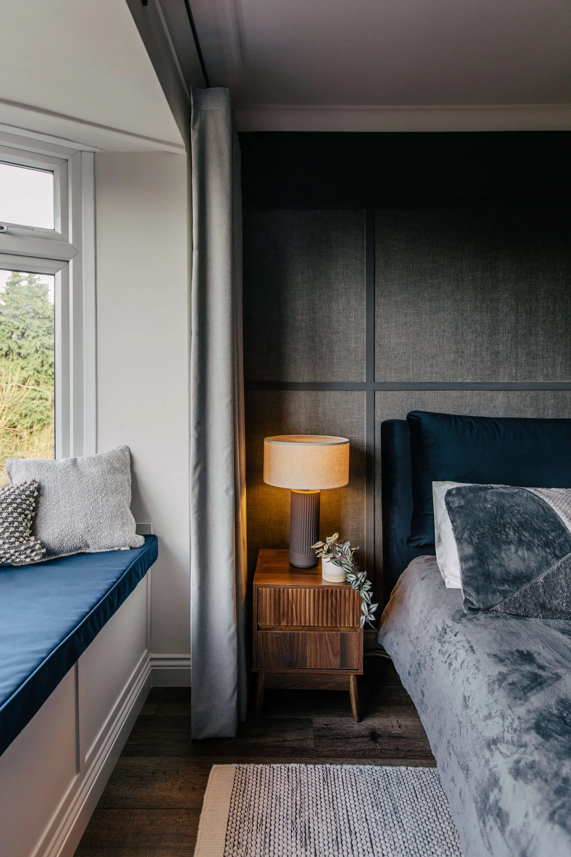 Interior bedroom with a window seat on the left with cushions, a nightstand with a lamp and plant beside a bed with dark and gray bedding, against a dark upholstered headboard and gray wall panels.