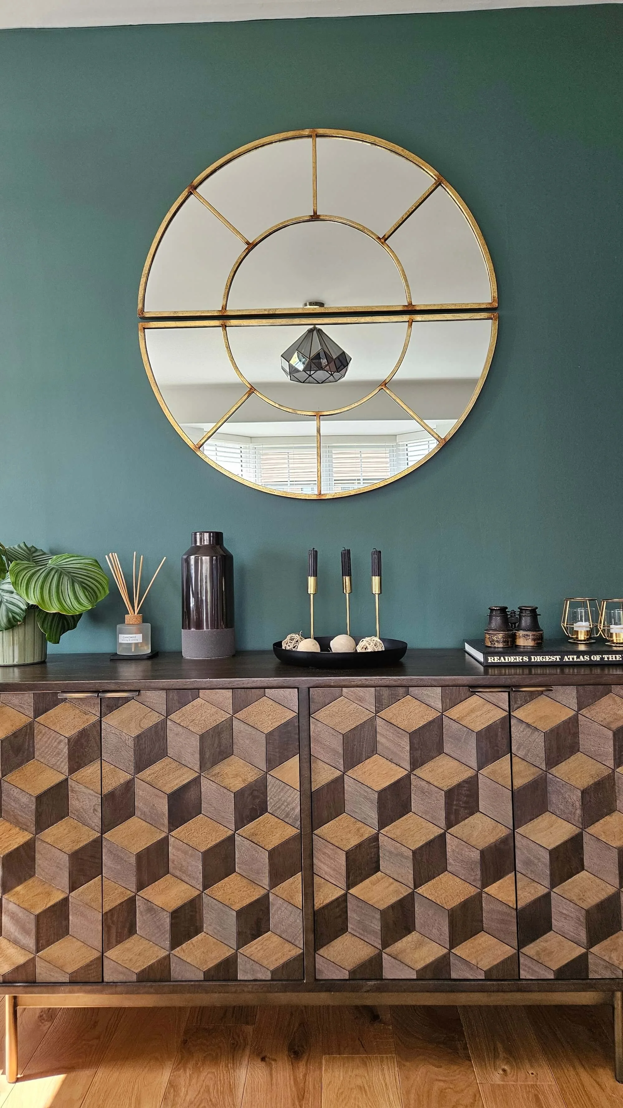 Decorative sideboard with geometric patterned doors, topped with a black dish holding candles, a black vase, a small plant, and glasses, against a green wall with a large round gold-framed mirror reflecting a ceiling light and windows.