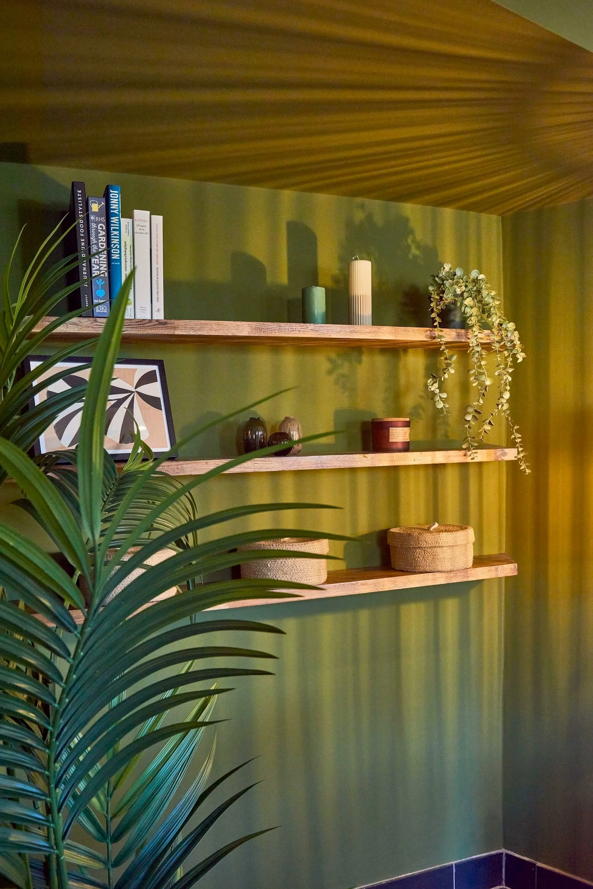 Green wall with three wooden shelves holding books, candles, and decorative items, with a potted plant in the foreground.