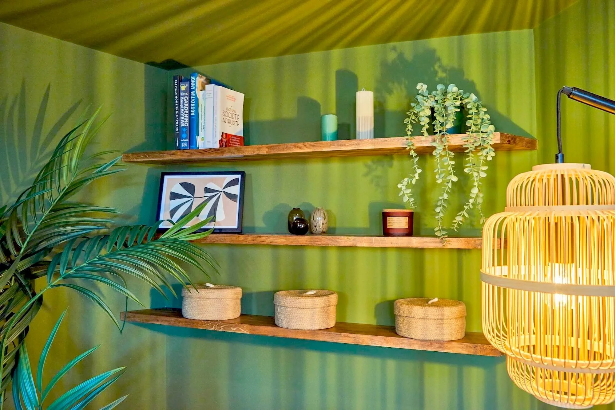Interior of a room with three wooden shelves on a bright green wall, decorated with books, framed artwork, small vases, candles, and trailing plant, adjacent to a large potted plant and a woven pendant lamp.