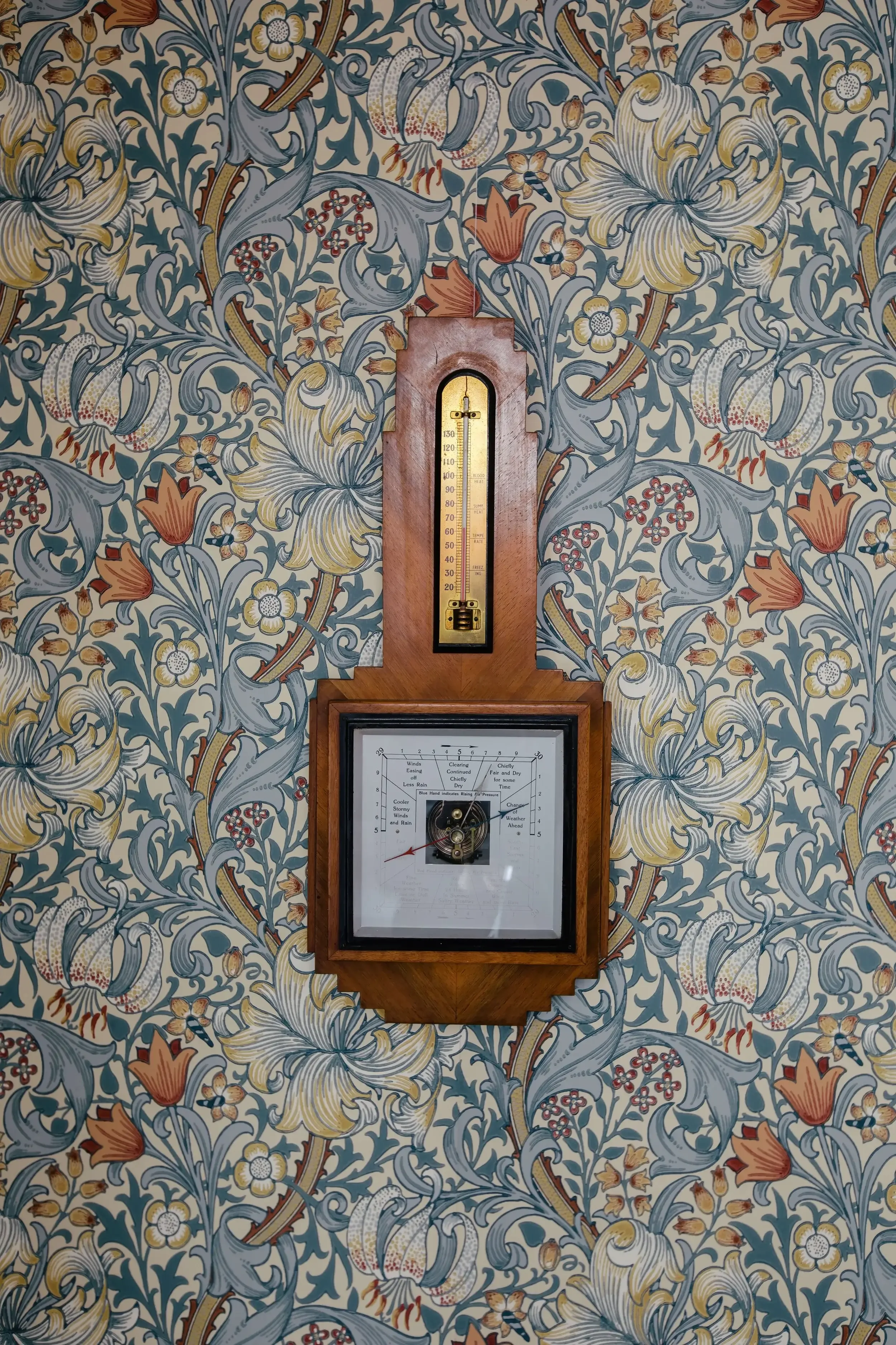 A vintage weather station with a thermometer and barometer mounted on a wall with floral wallpaper.