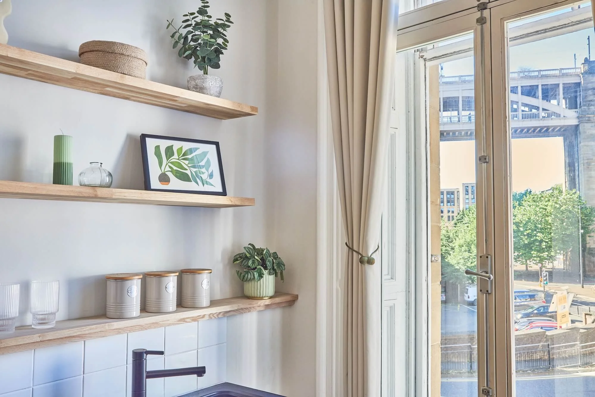 Interior of a kitchen with wooden open shelves decorated with plants, jars, and a framed botanical print, near a window with beige curtains showing a cityscape and a bridge outside.