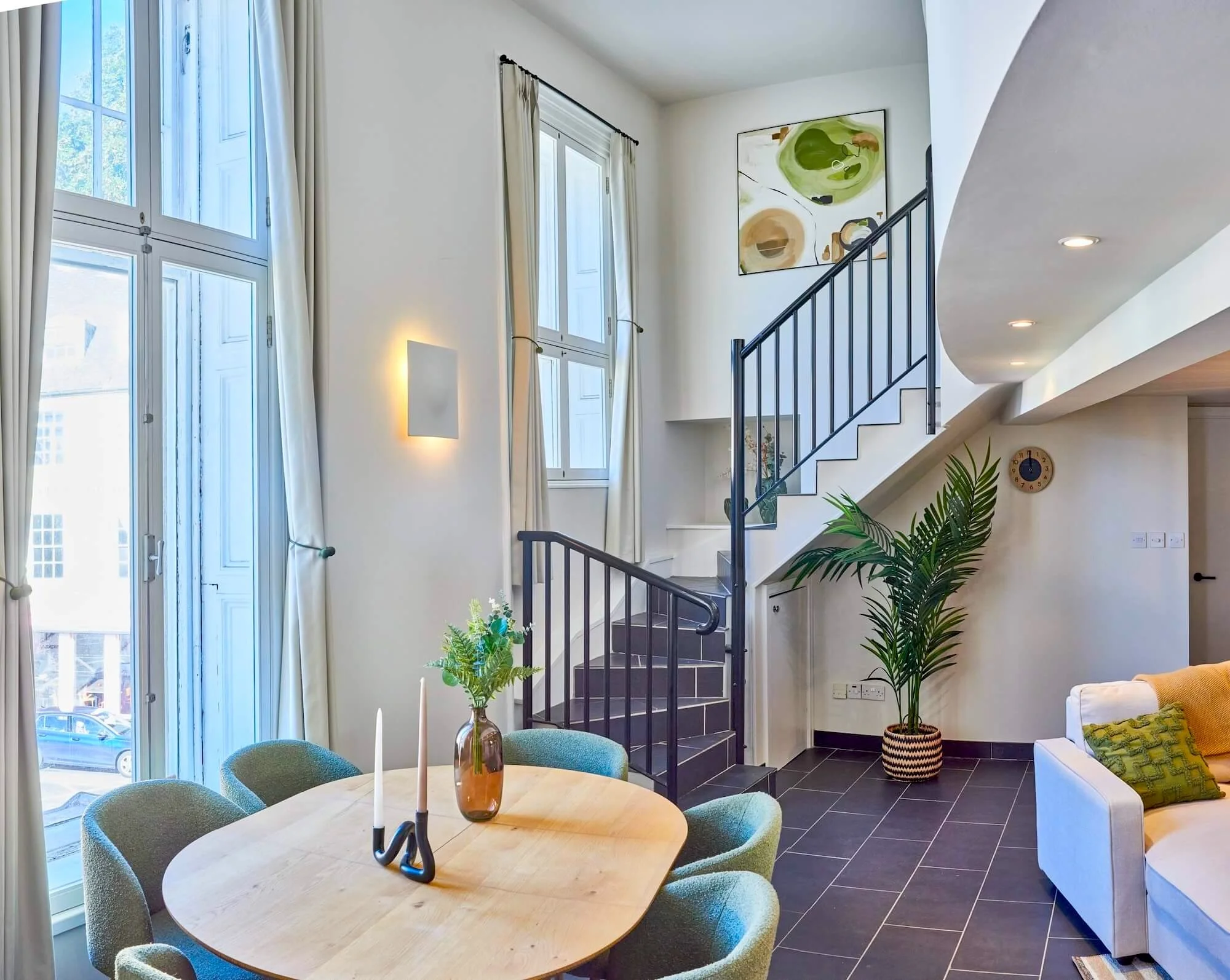 Bright living room with large windows, white curtains, a round wooden dining table with green chairs, a white sofa with a green throw pillow, a potted plant, and modern art on the wall.