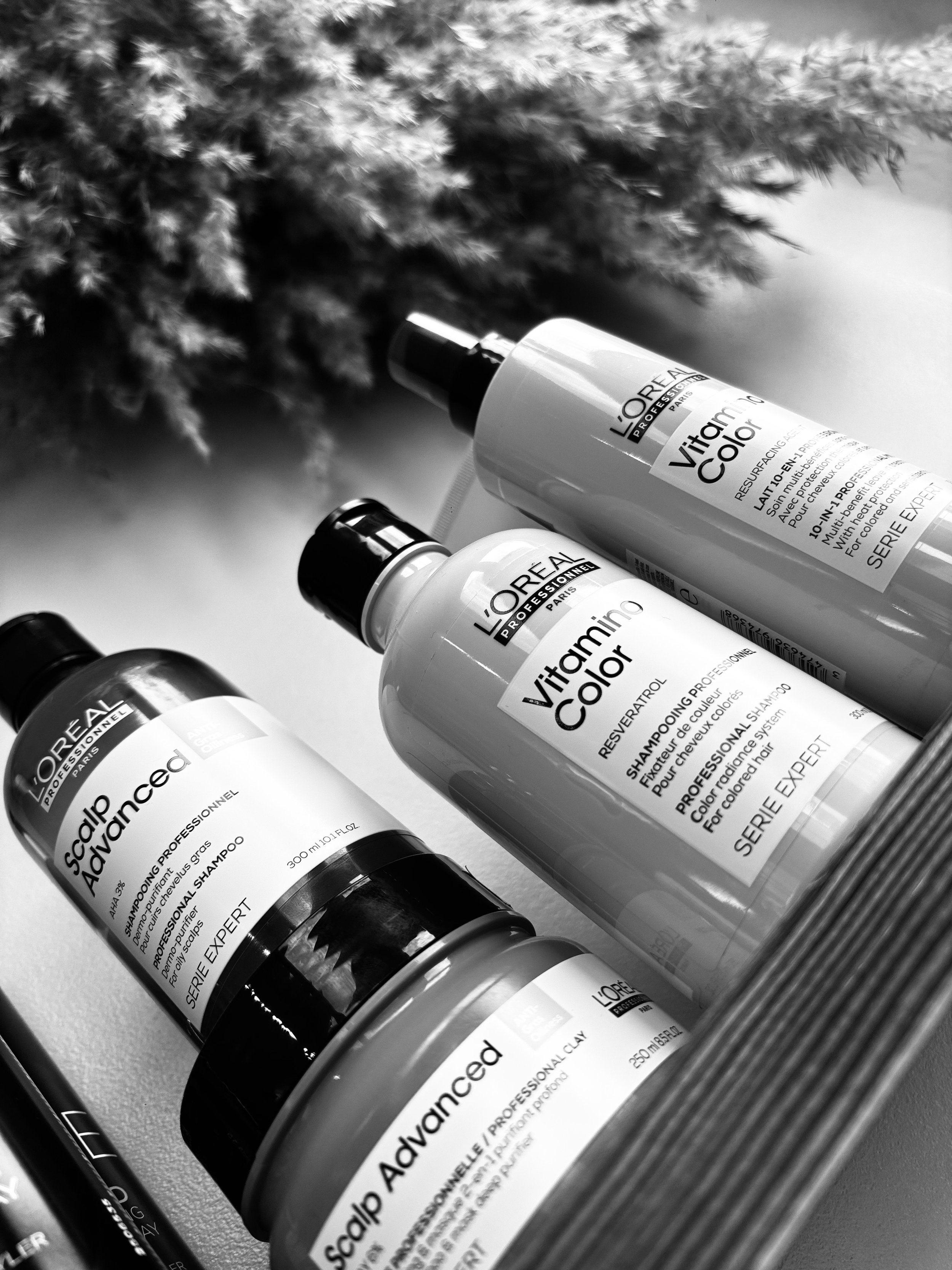 Black and white photo of three L'Oréal Professionnel Vitamino Color hair care bottles on a surface with blurred background of a plant.