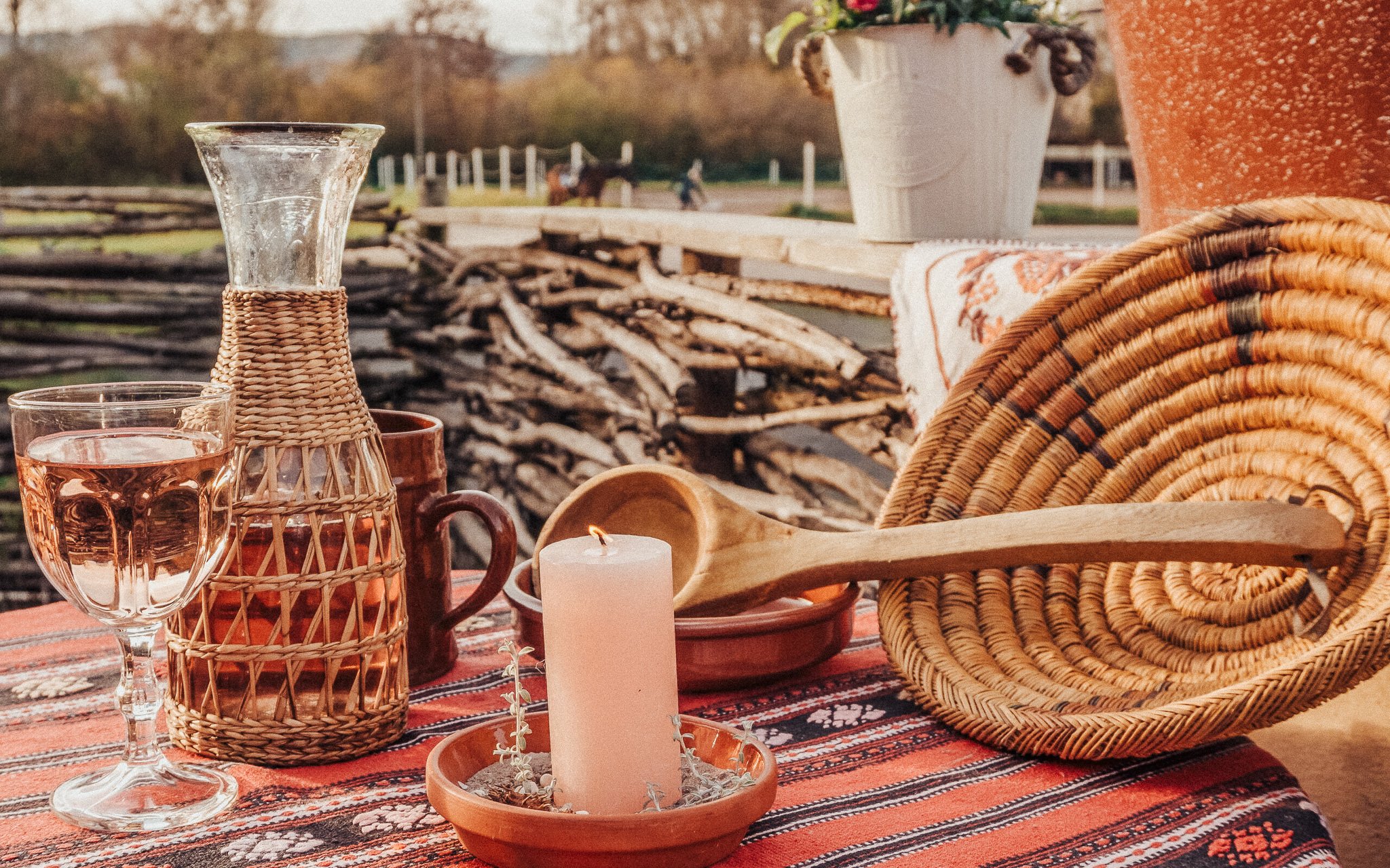 Ein Tisch mit Kerze, Weinglas, Wasserkrug mit Keramikummantelung, Tasse, Holzlöffel, Korbuntersetzer und Topfpflanze, im Freien mit Holzstapeln und Pferden im Hintergrund.