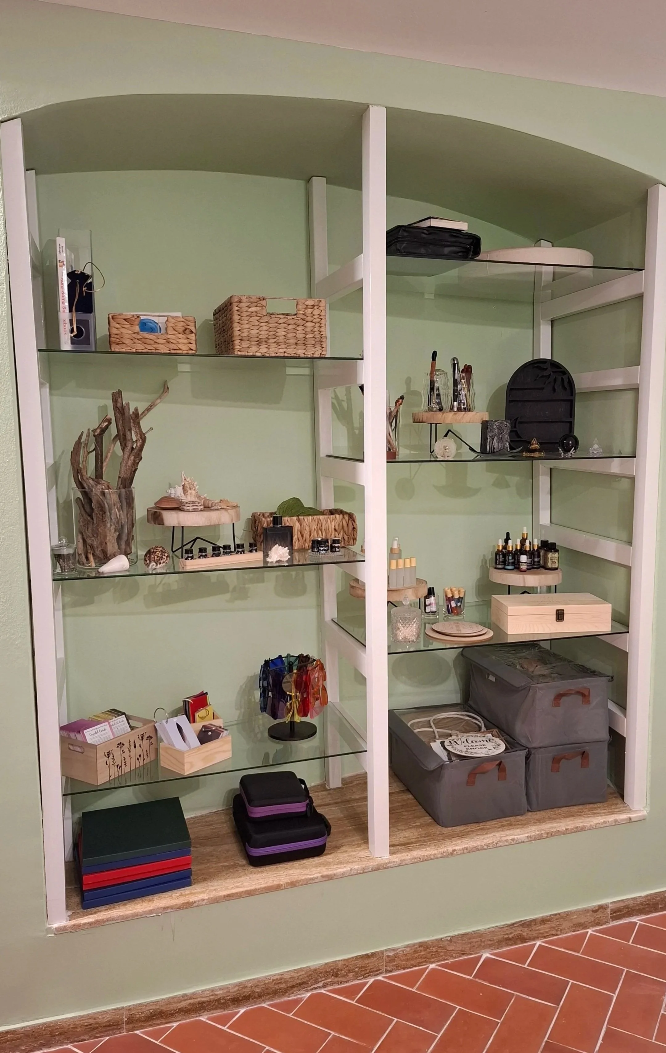 Open wooden and glass shelving unit with various objects including baskets, decorative items, essential oils, storage boxes, and small containers, against a green wall on a tiled floor.