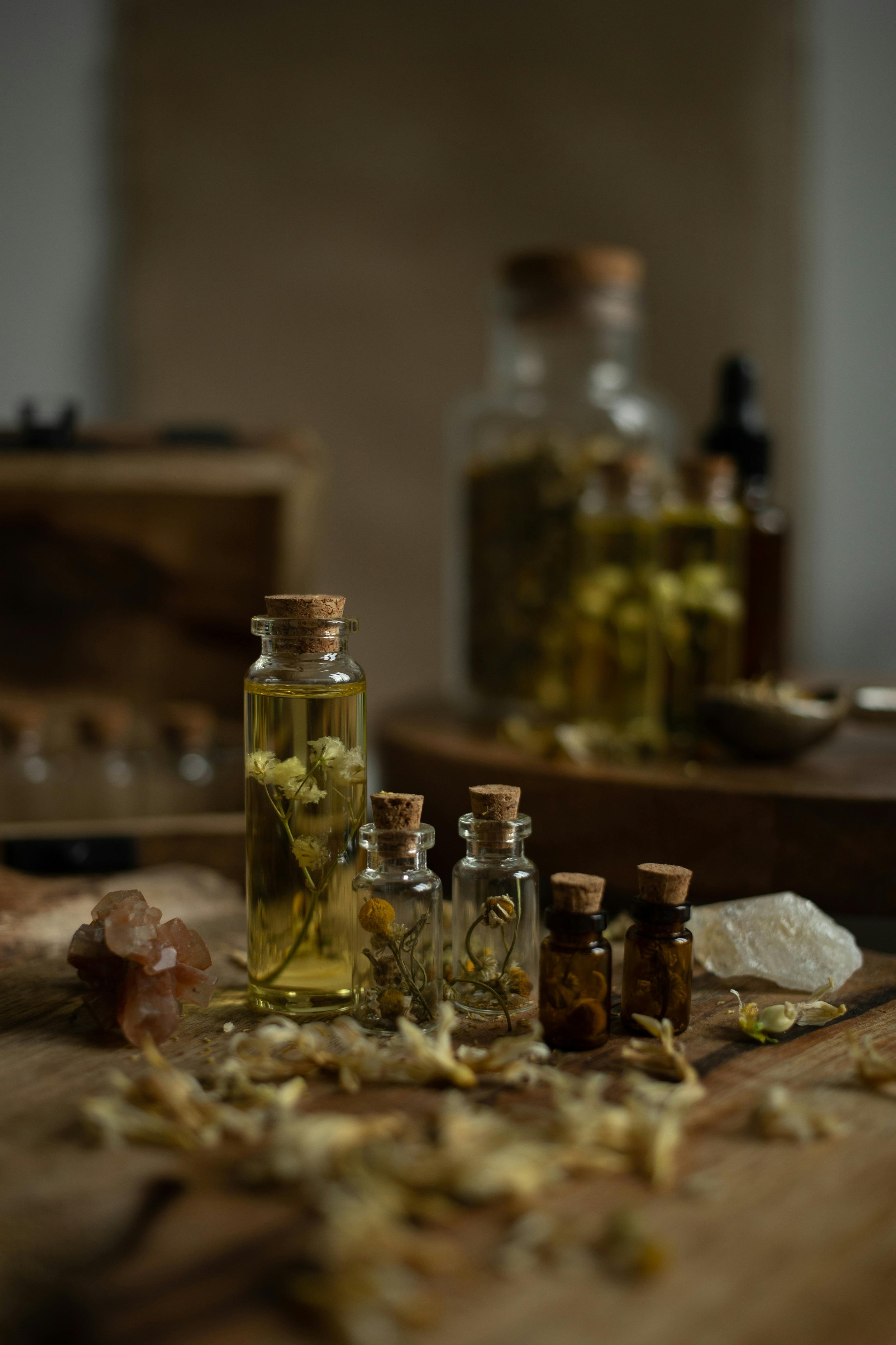 Small glass bottles and jars with cork stoppers containing dried flowers and herbs, on a wooden surface, with larger jars and bottles in the background.