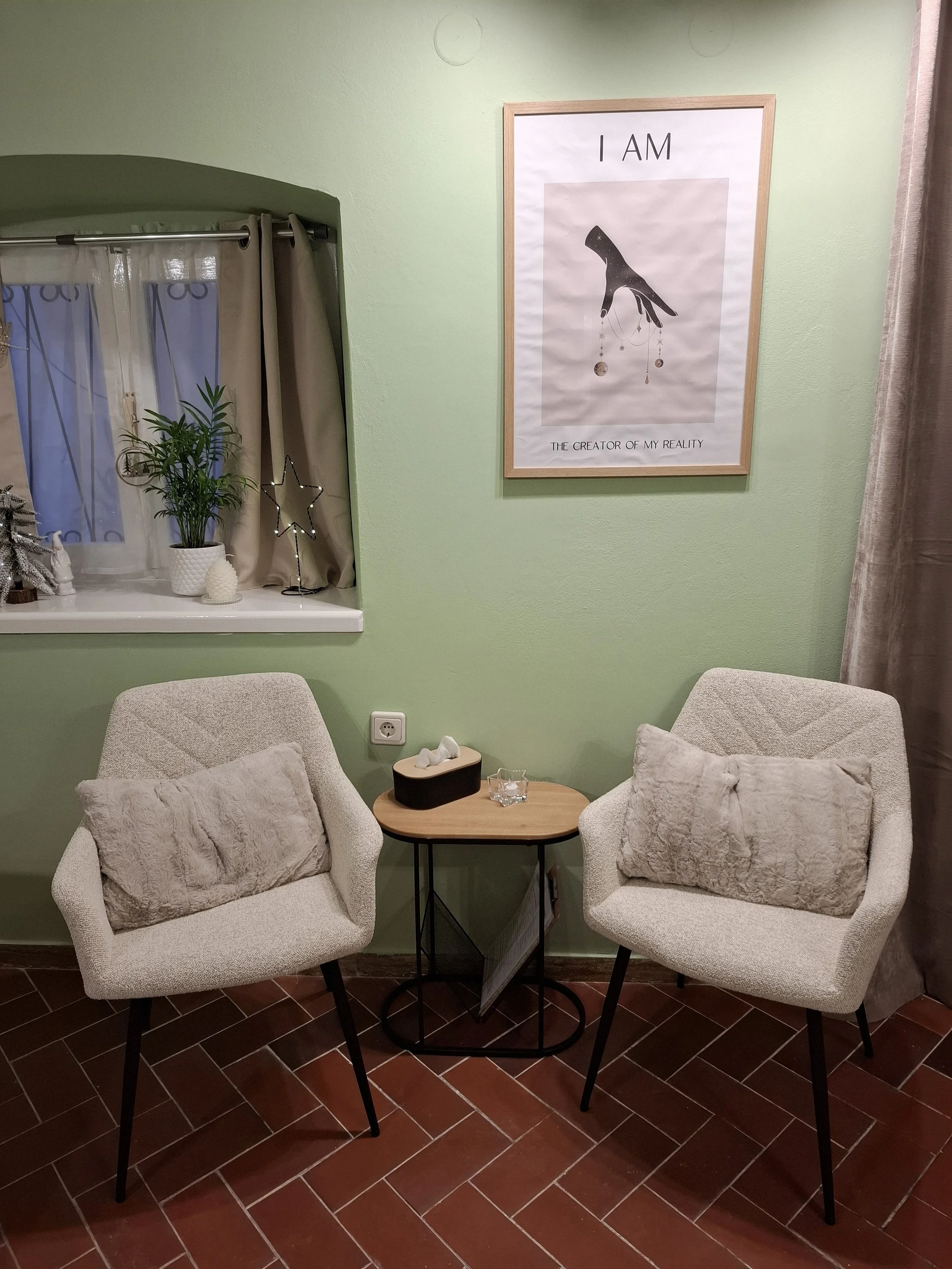 A cozy corner with two beige armchairs with fluffy pillows, a small wooden side table, a window with plants and decorations, and a framed artwork on a green wall that says 'I AM THE CREATOR OF MY REALITY' with a black and white abstract hand design.