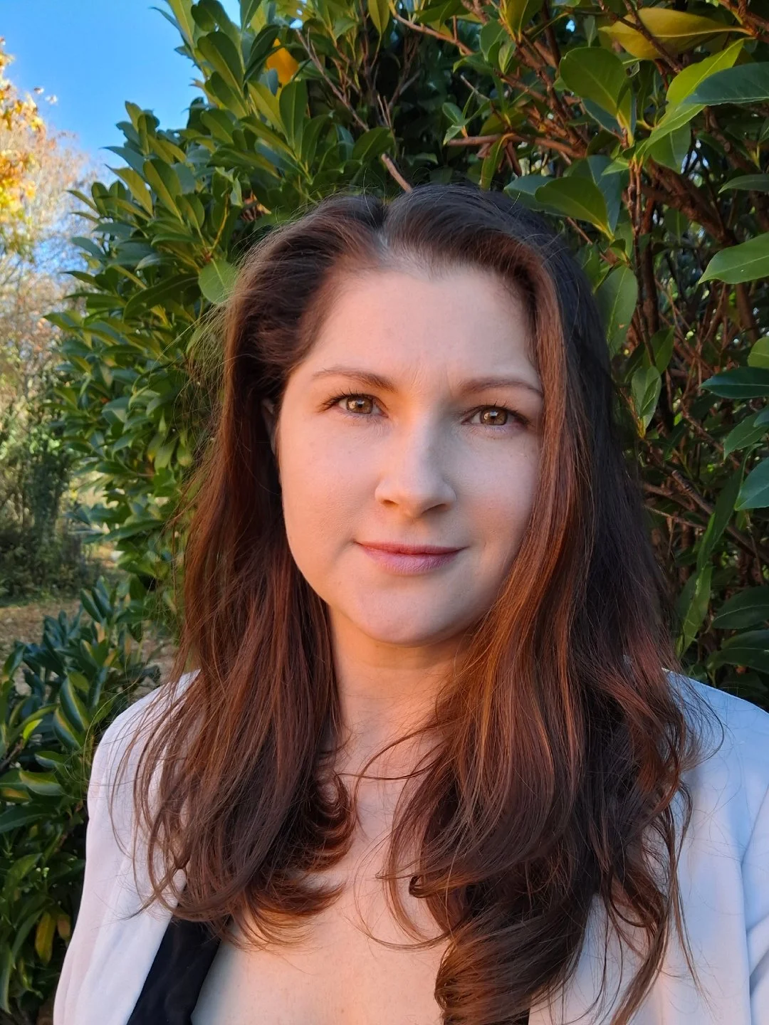 A woman with long brown hair standing outdoors in front of green bushes, smiling softly at the camera during golden hour sunlight.