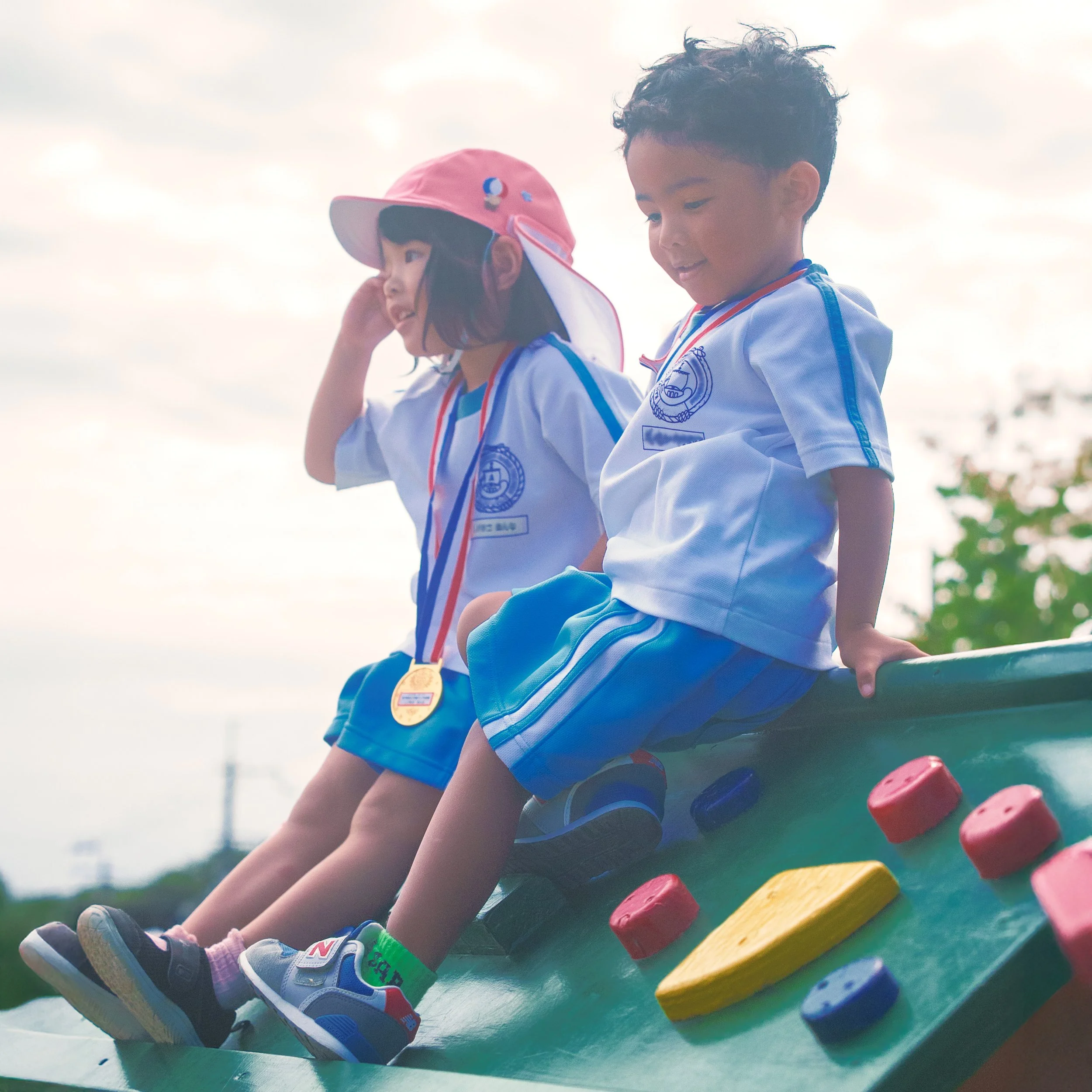 公園の遊具の上で遊ぶ小さな男の子と女の子。男の子は白いスポーツユニフォームを着て座っており、女の子はキャップをかぶって同じ制服を着ている。