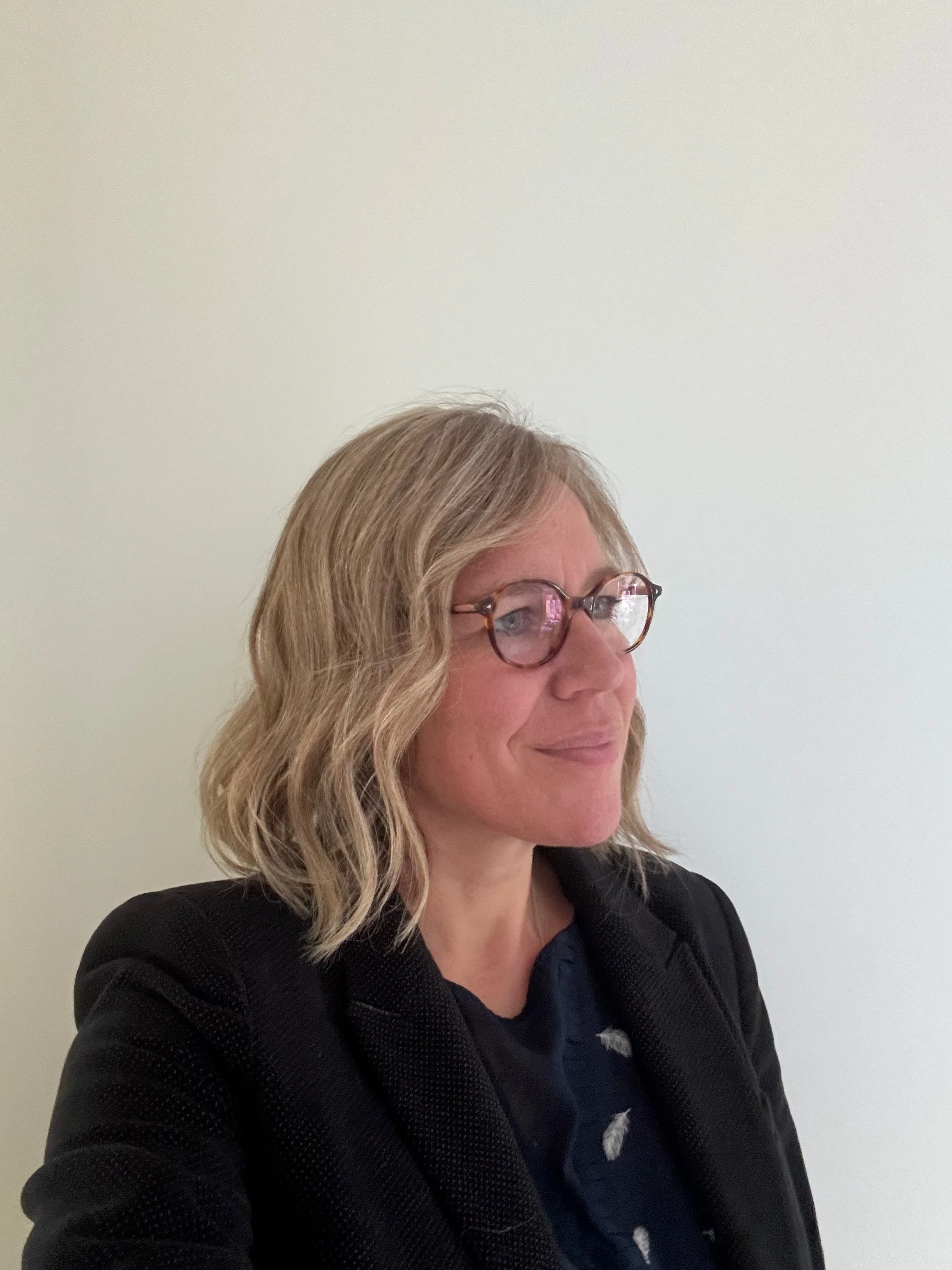 A photo of Alix who is a woman with shoulder-length wavy blonde-grey hair wearing glasses, a dark patterned shirt, and a black blazer standing against a plain white wall.