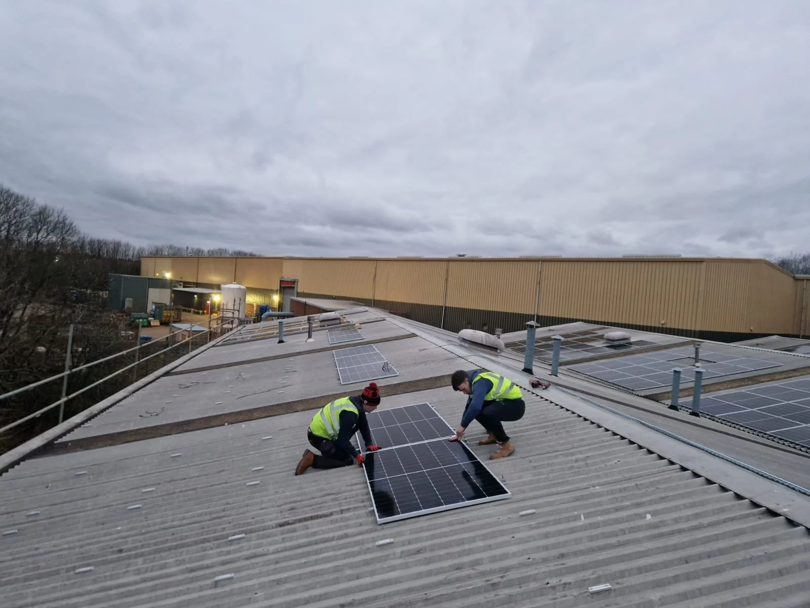Two workers in safety vests installing solar panels on a large commercial building's metal roof in cloudy weather.