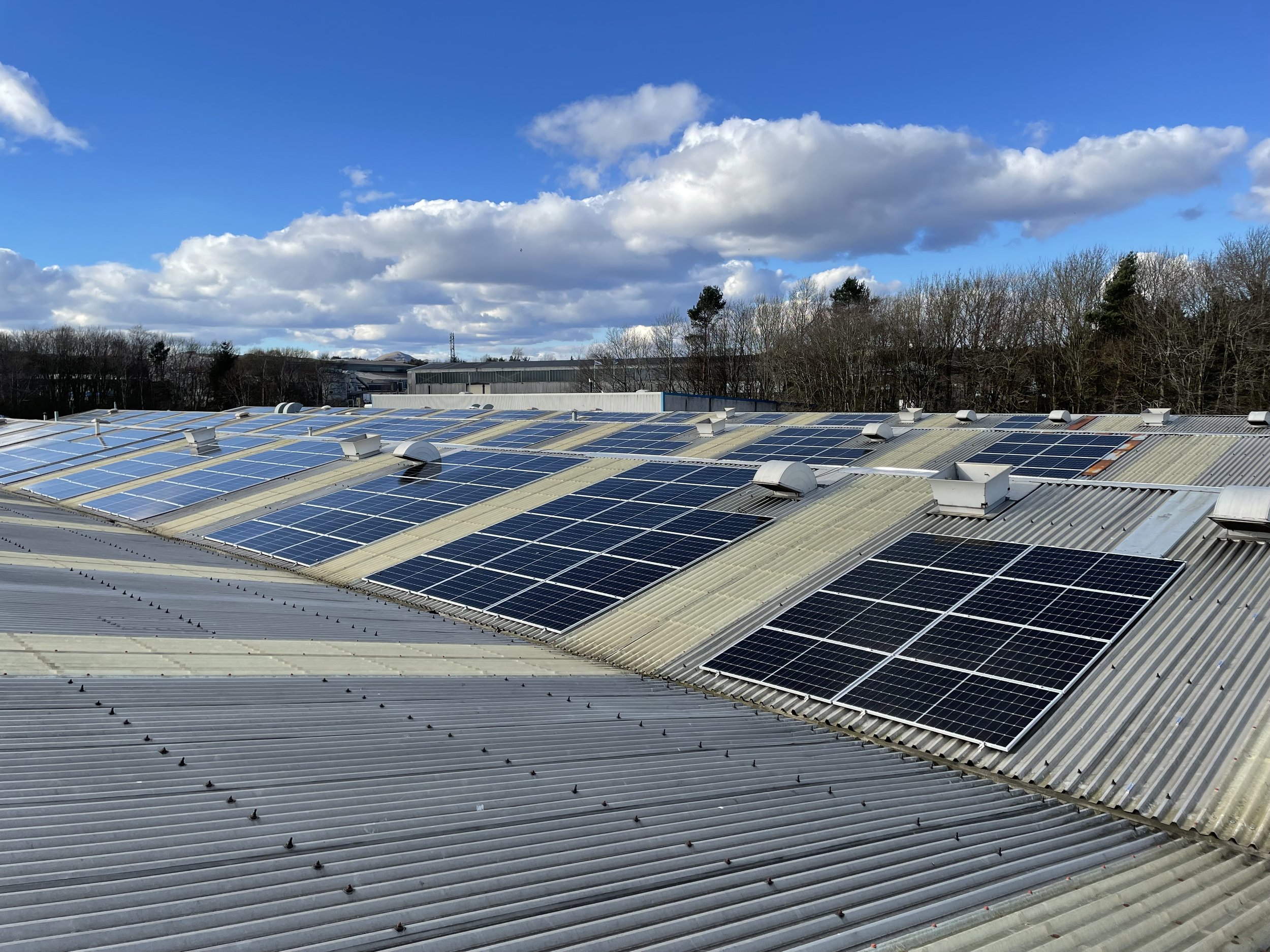 A rooftop covered with numerous solar panels under a partly cloudy sky with trees in the background.