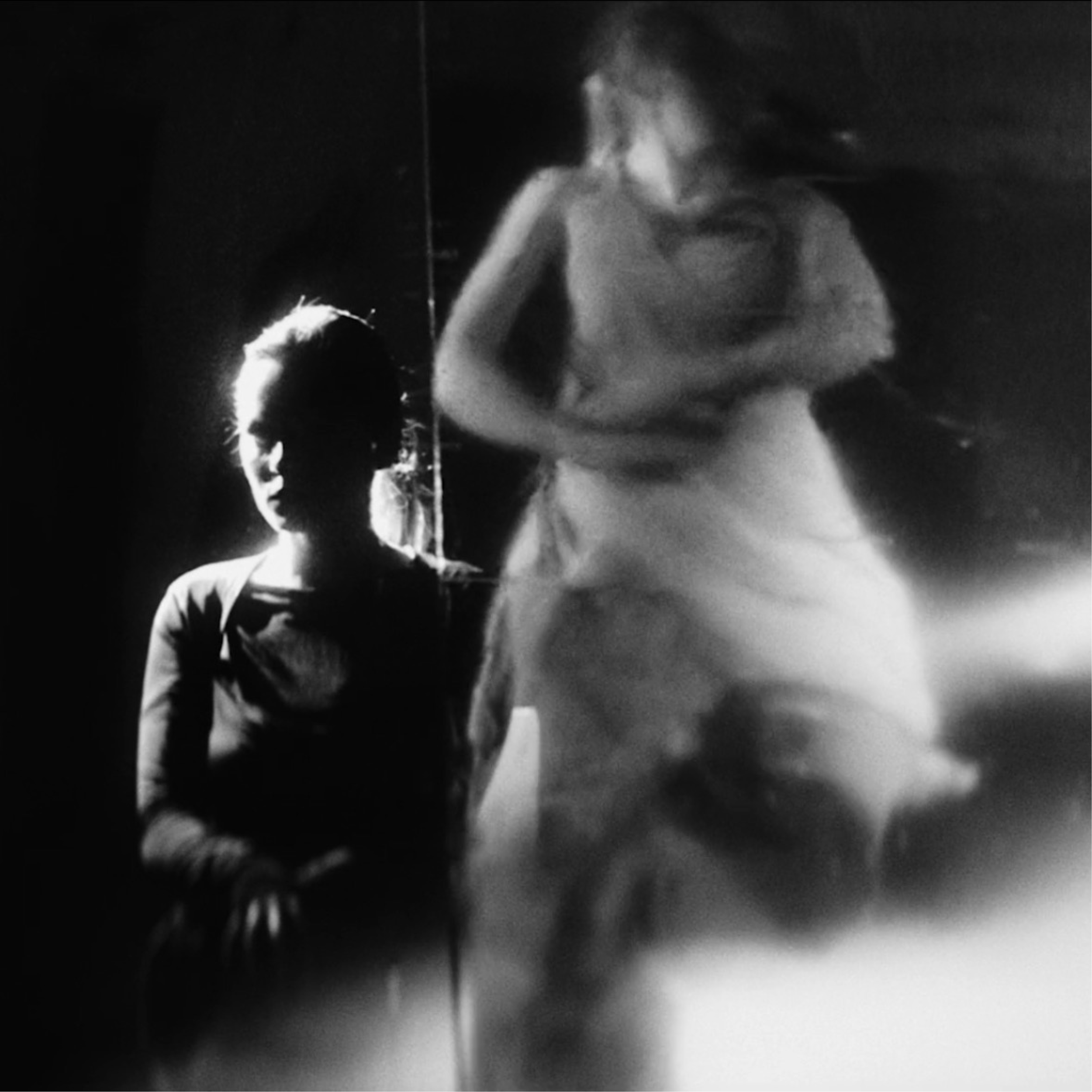 Black and white photo of two women, one sitting and one standing, with dramatic lighting casting shadows. The seated woman has her face partially illuminated, and the standing woman has her head turned with her hair flowing.