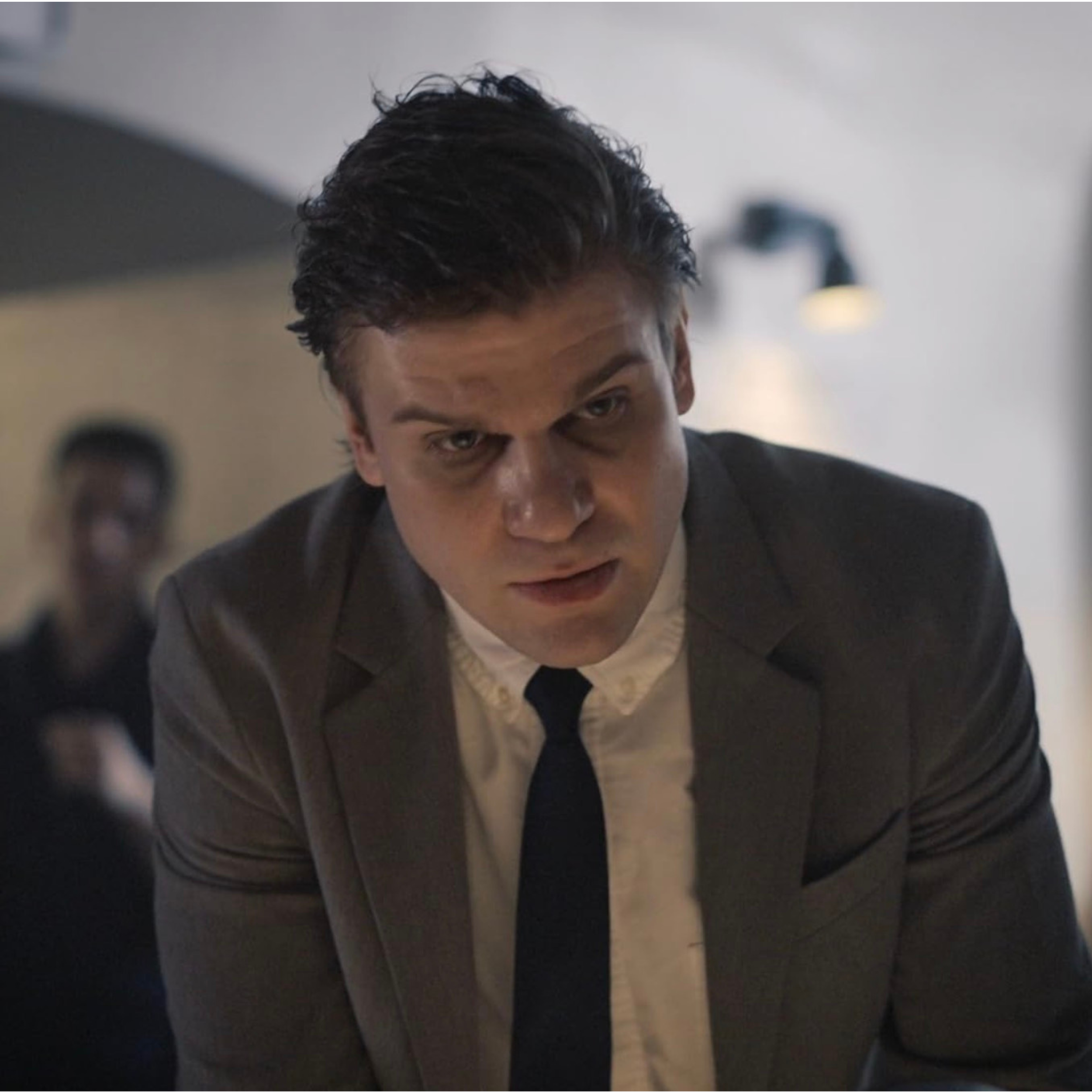 A man in a gray suit and black tie leaning forward, with a serious or angry expression on his face. In the background, there is a blurred person and a wall lamp.
