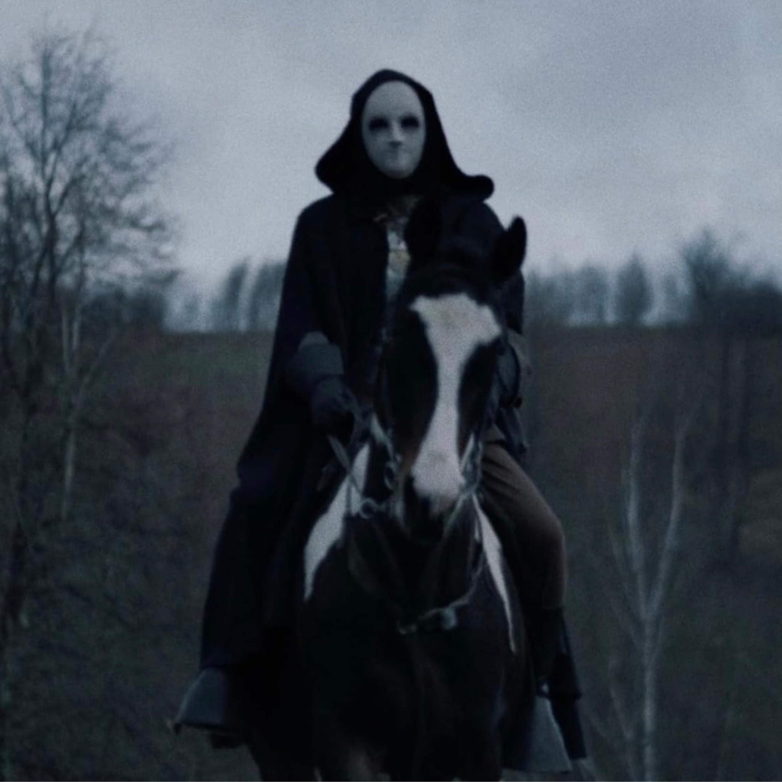 Person in a mask riding a horse outdoors at dusk or dawn, with leafless trees in the background.
