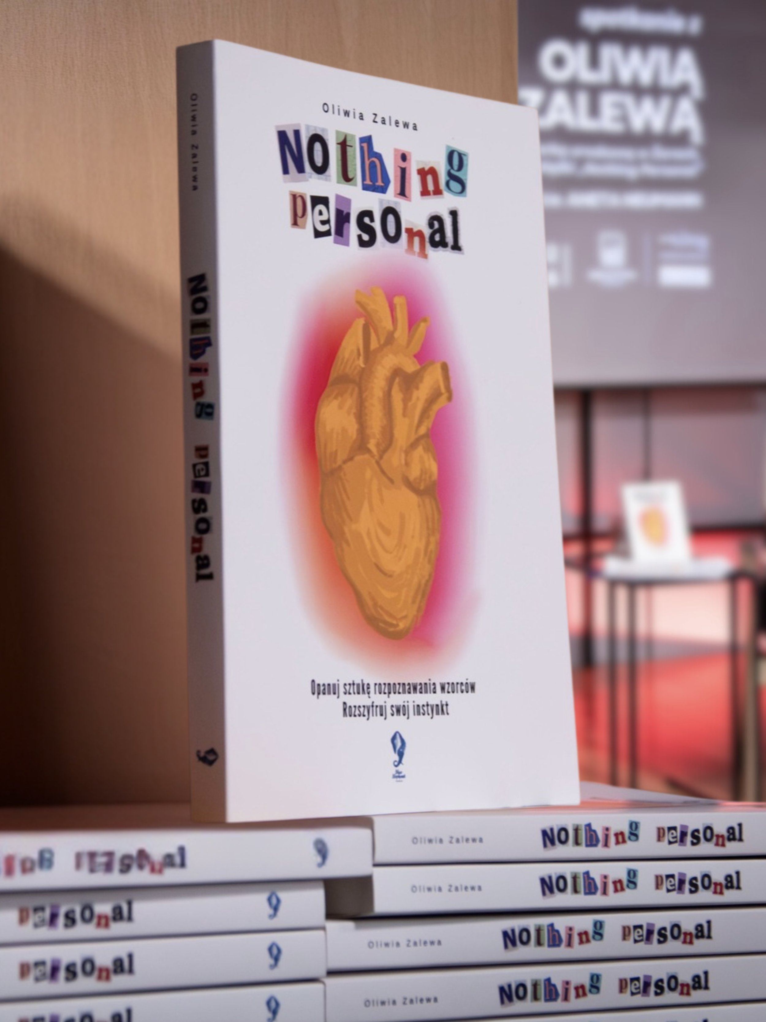 A book titled 'Nothing Personal' by Oliwia Zalewa displayed on a table, with multiple copies stacked in front. The cover features an illustration of a human heart and colorful, cut-out style text.