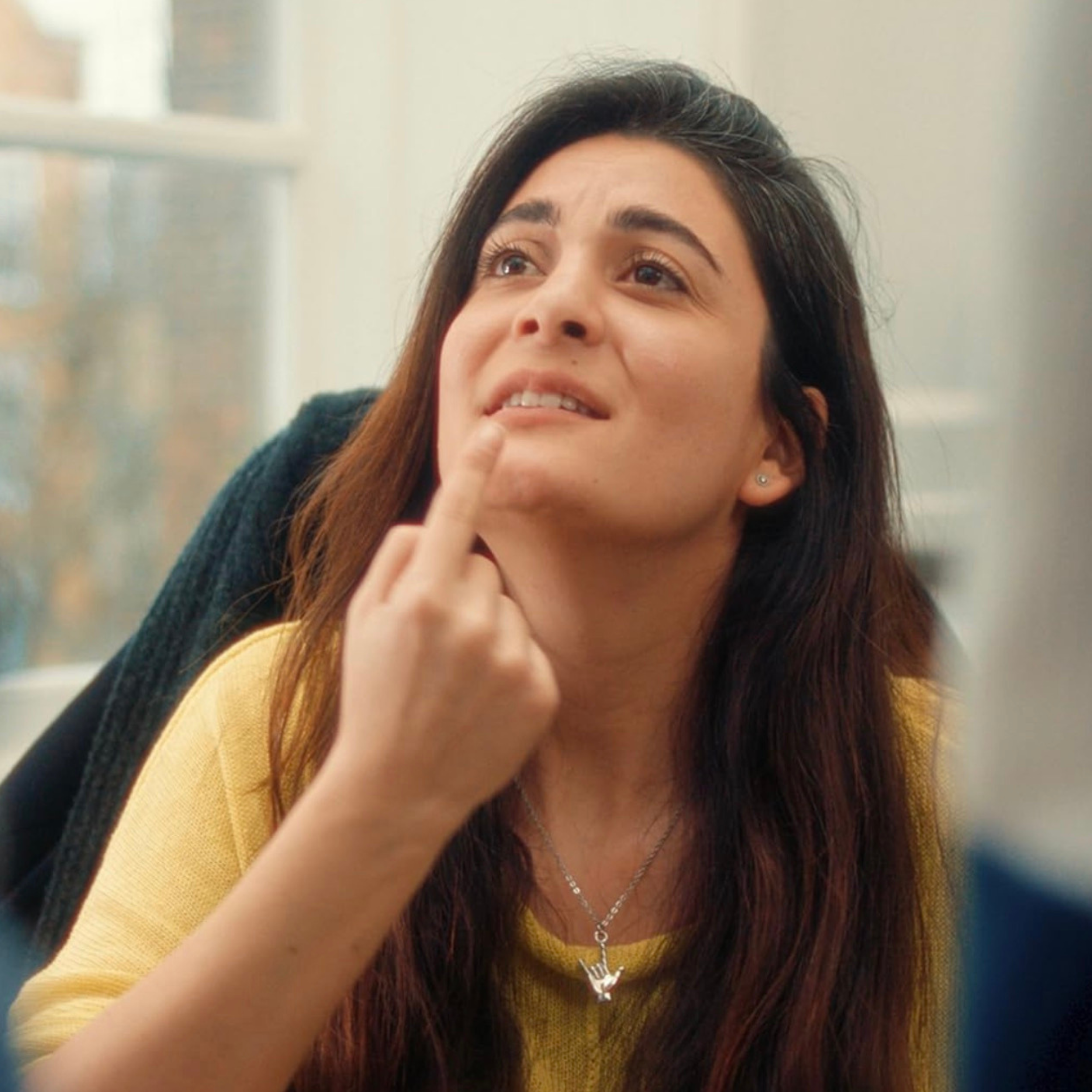 A woman with long dark hair, wearing a yellow top and a silver necklace, touching her chin thoughtfully in a bright room.