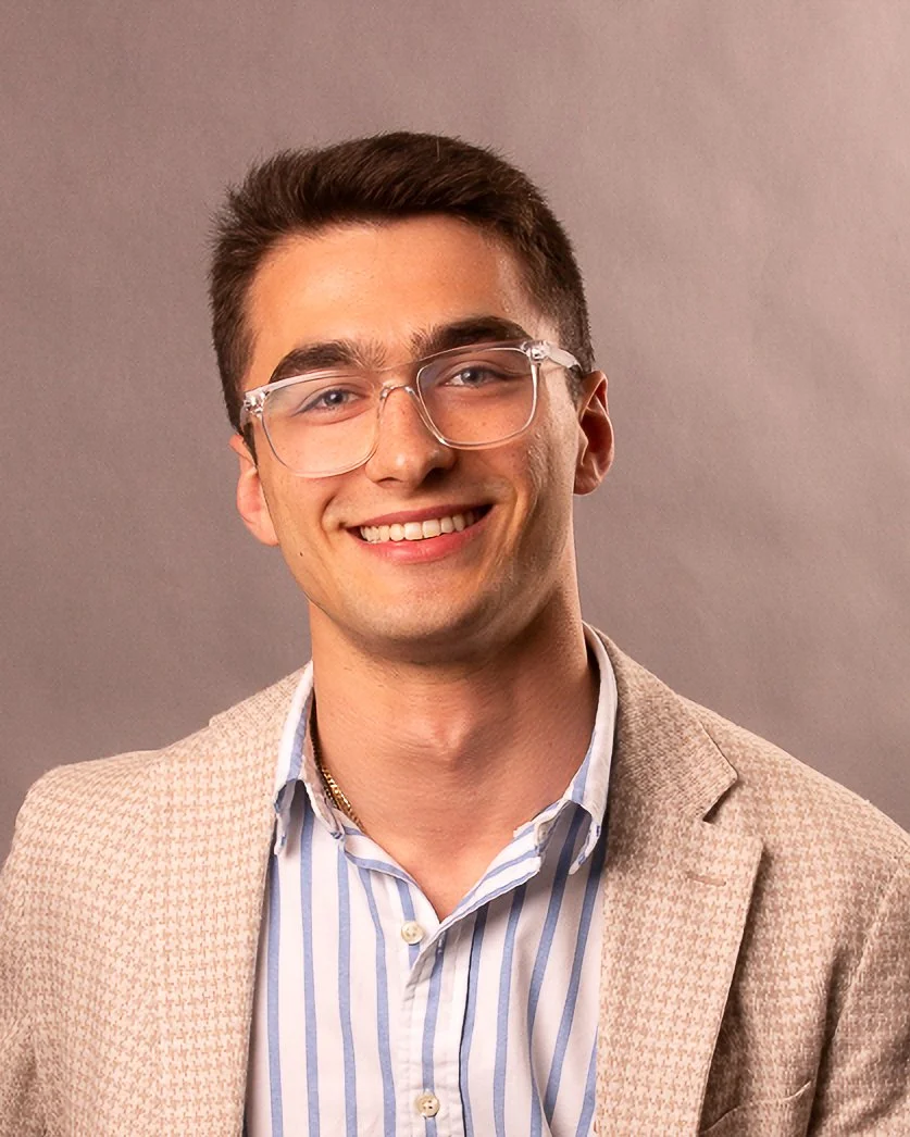 A young man with short dark hair, wearing clear glasses, a beige blazer, and a striped shirt, smiling at the camera against a gray background.