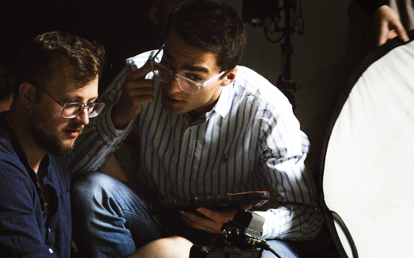 Two men working closely together on a project, wearing glasses, with one holding a tablet and the other looking at the screen, in a dimly lit environment.