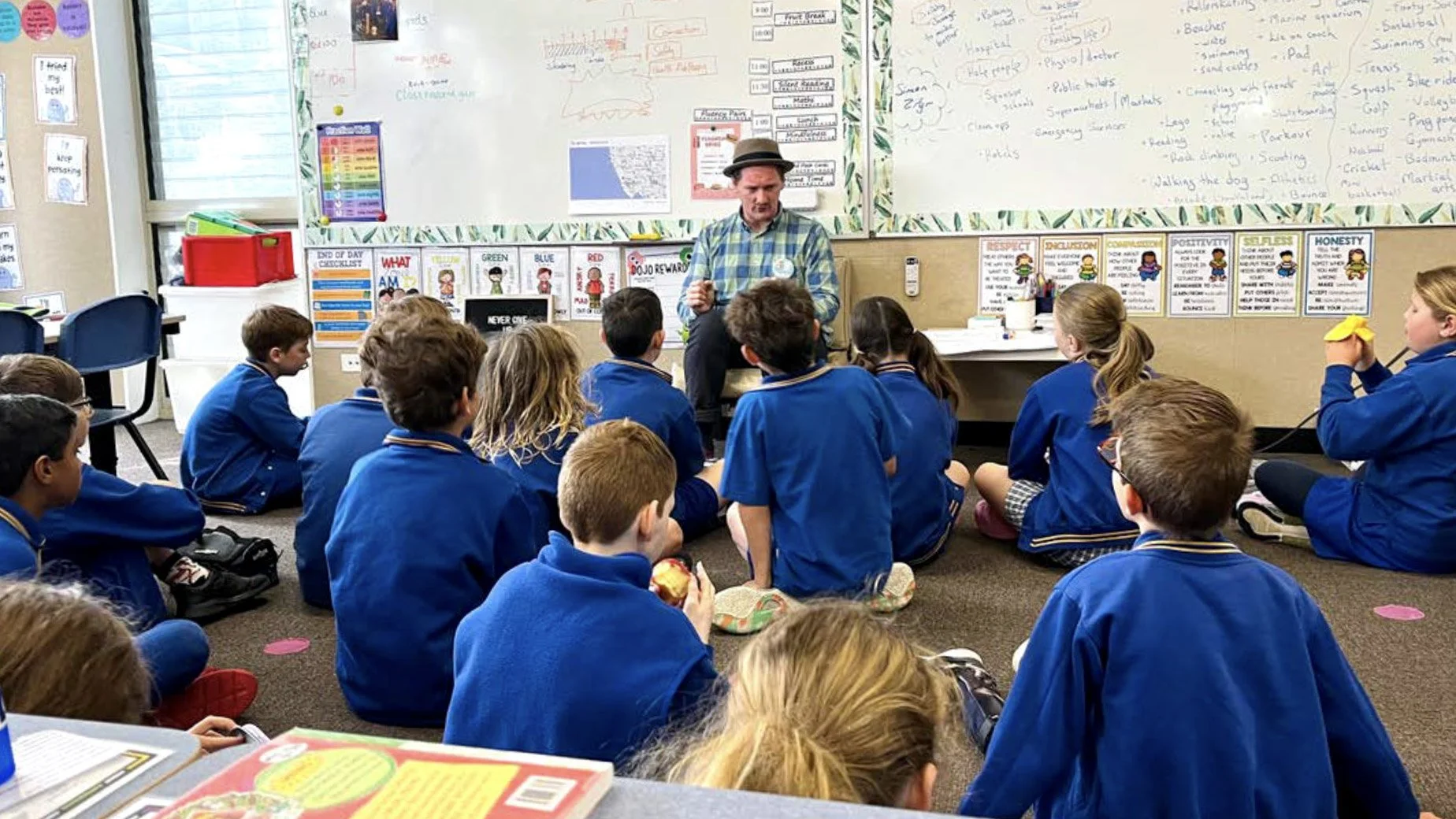 A facilitator sitting in front of a whiteboard speaks to a primary school class