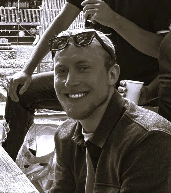 A smiling young man with sunglasses on his head sitting outdoors at a table with other people nearby.