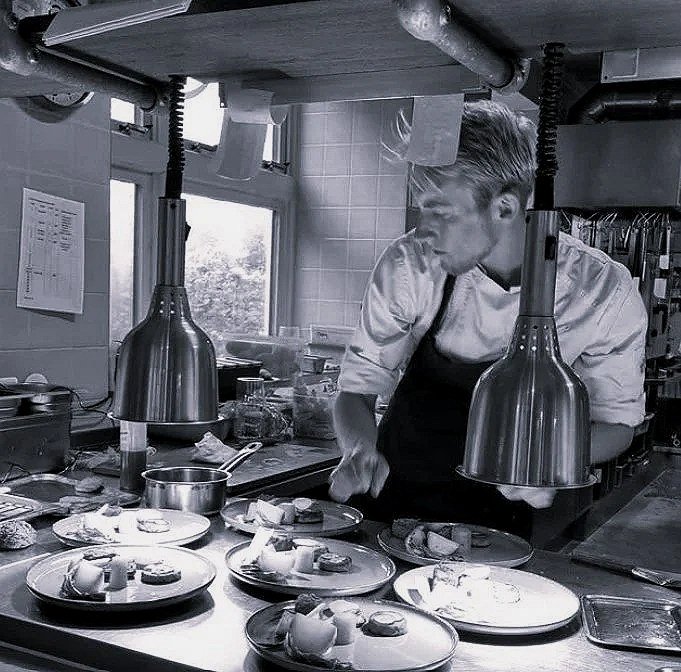 A chef in a professional kitchen preparing plates with dishes of food, under two hanging heat lamps.
