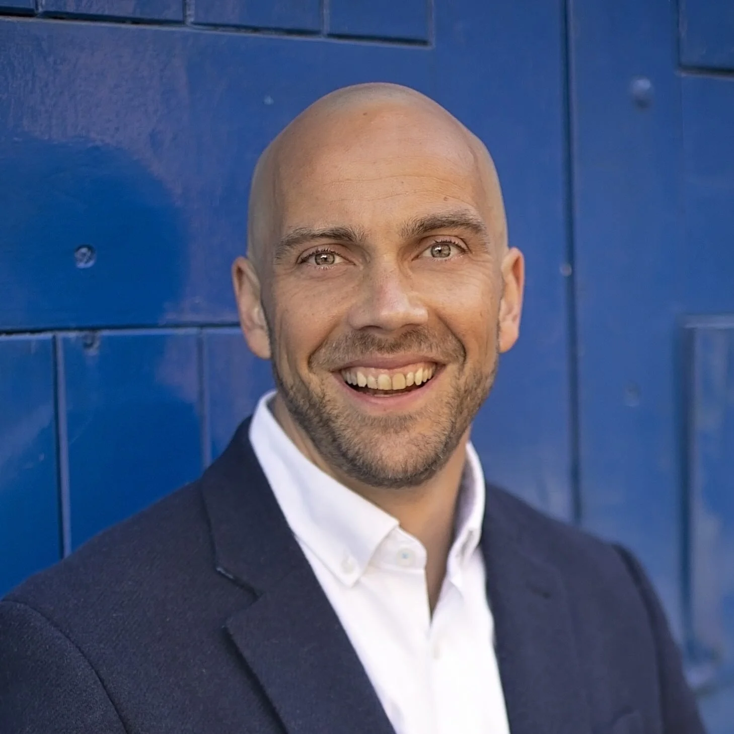 Portrait of a smiling man with a shaved head and light eyes, wearing a dark suit jacket and white shirt, standing in front of a blue wooden wall.