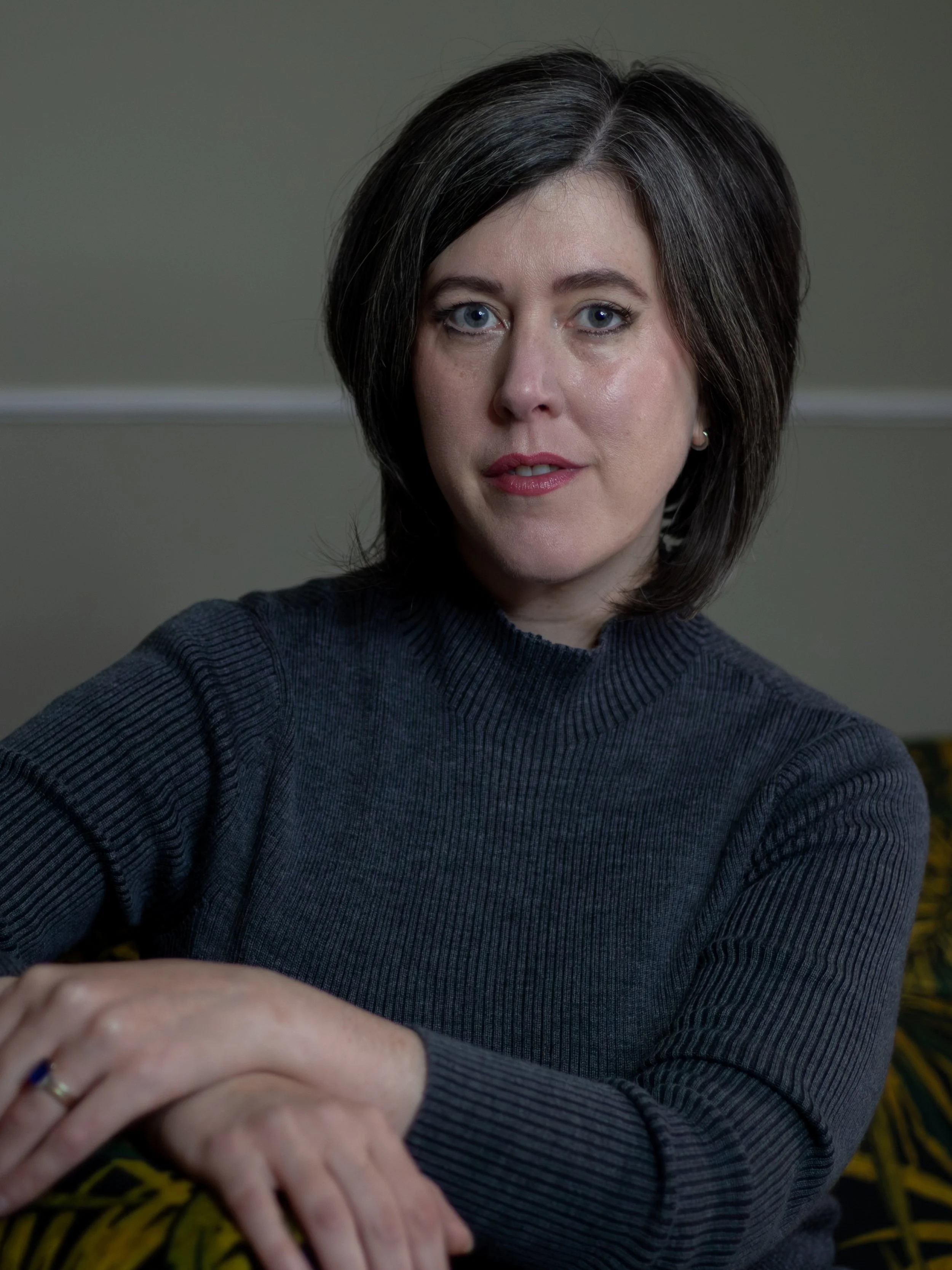 Portrait of a woman with dark brown hair and blue eyes wearing a dark gray ribbed sweater, sitting with her arms crossed on a patterned yellow and black surface, in front of a plain gray wall.