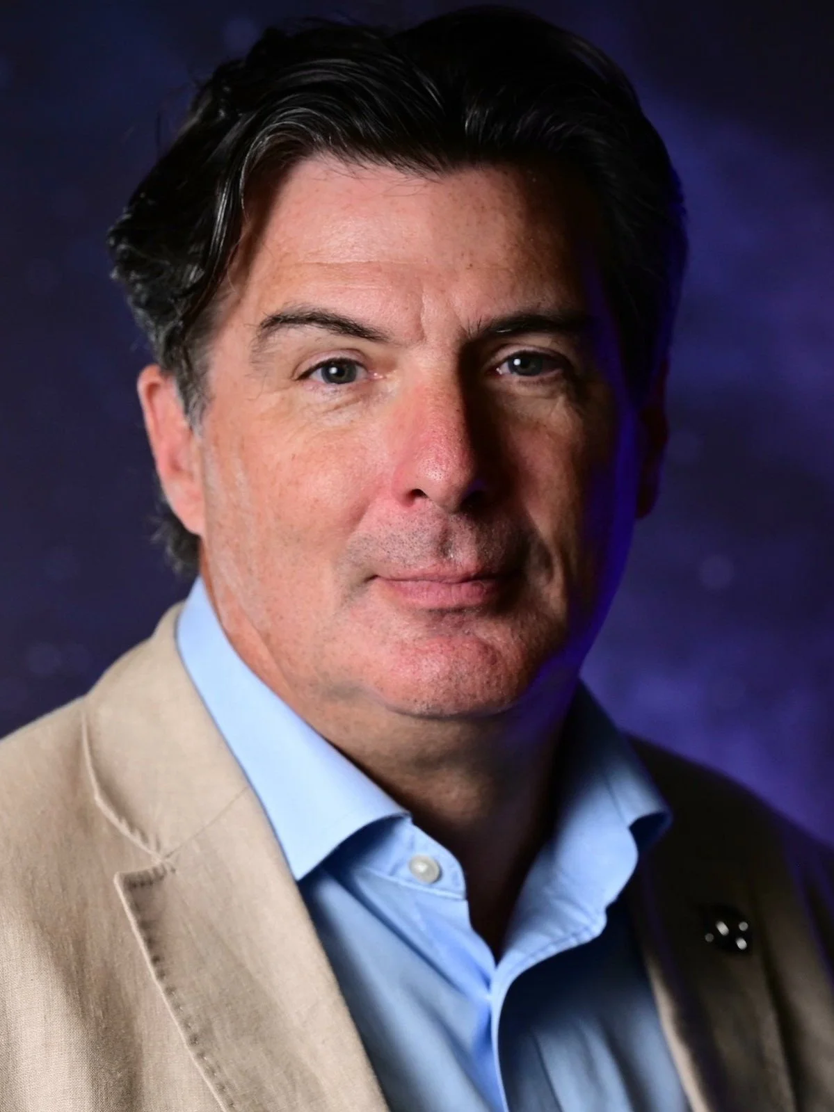 Close-up portrait of a man with dark, wavy hair, wearing a light blue shirt and beige blazer, against a dark, blurred background.