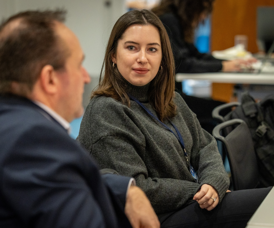 A woman with brown hair wearing a gray sweater and a lanyard, looking at a man with blurred face in a conference room or classroom setting.