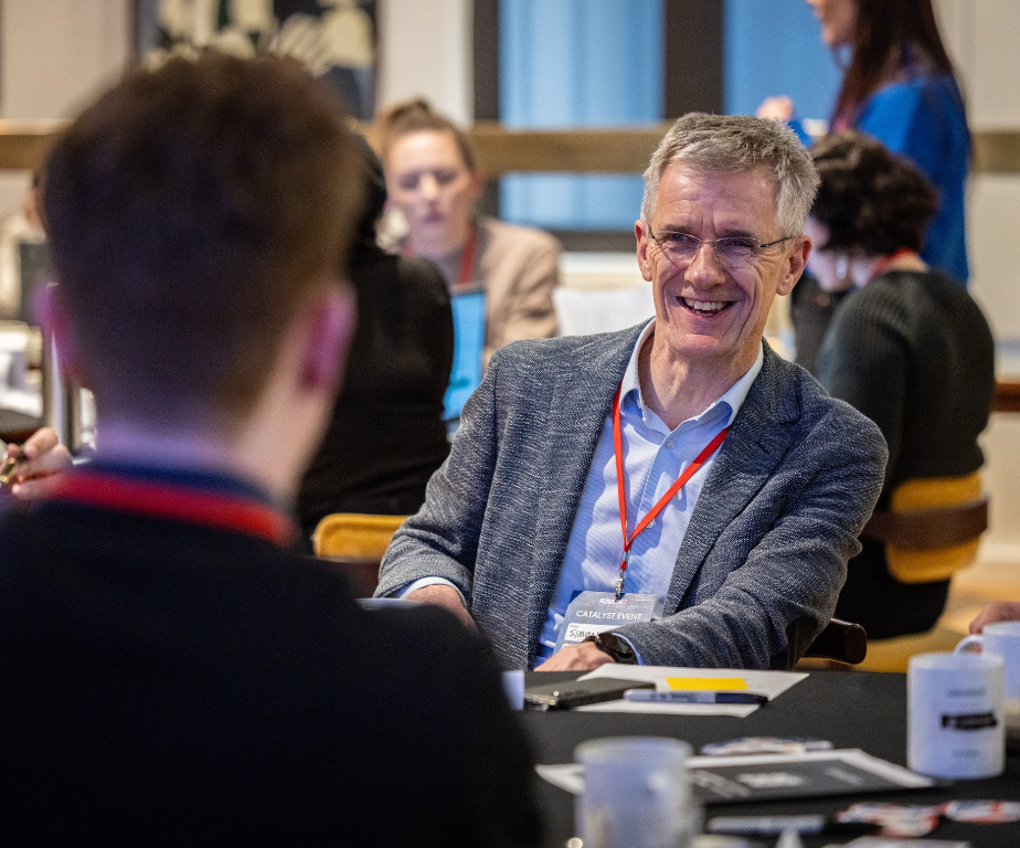 A man with gray hair, glasses, and a gray blazer smiling and talking to someone at a conference table with other attendees in the background.